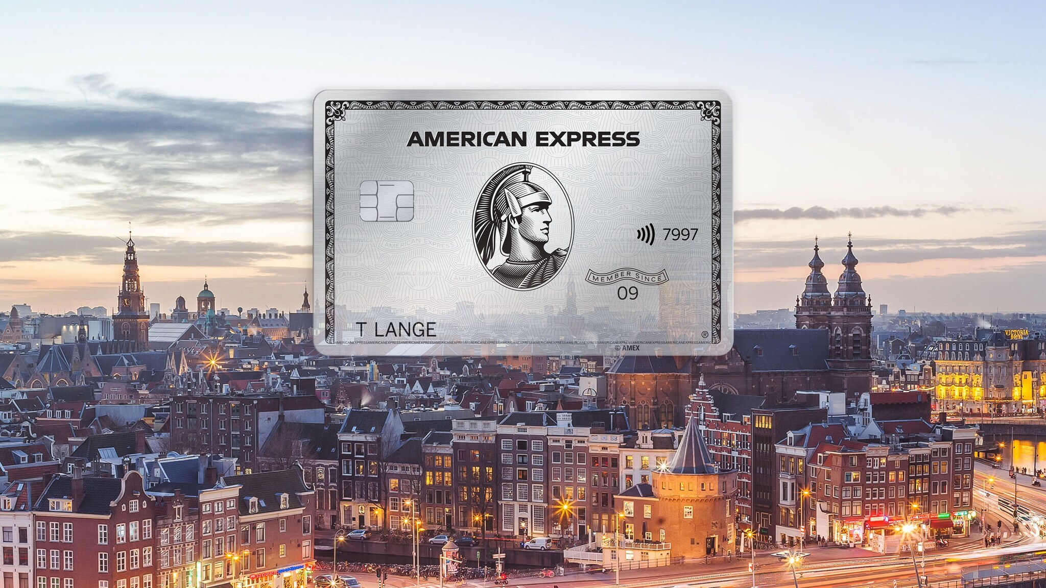 Fotocollage mit Freisteller einer silbernen Kreditkarte von American Express vor dem Stadtpanorama von Amsterdam in der Abenddämmerung.