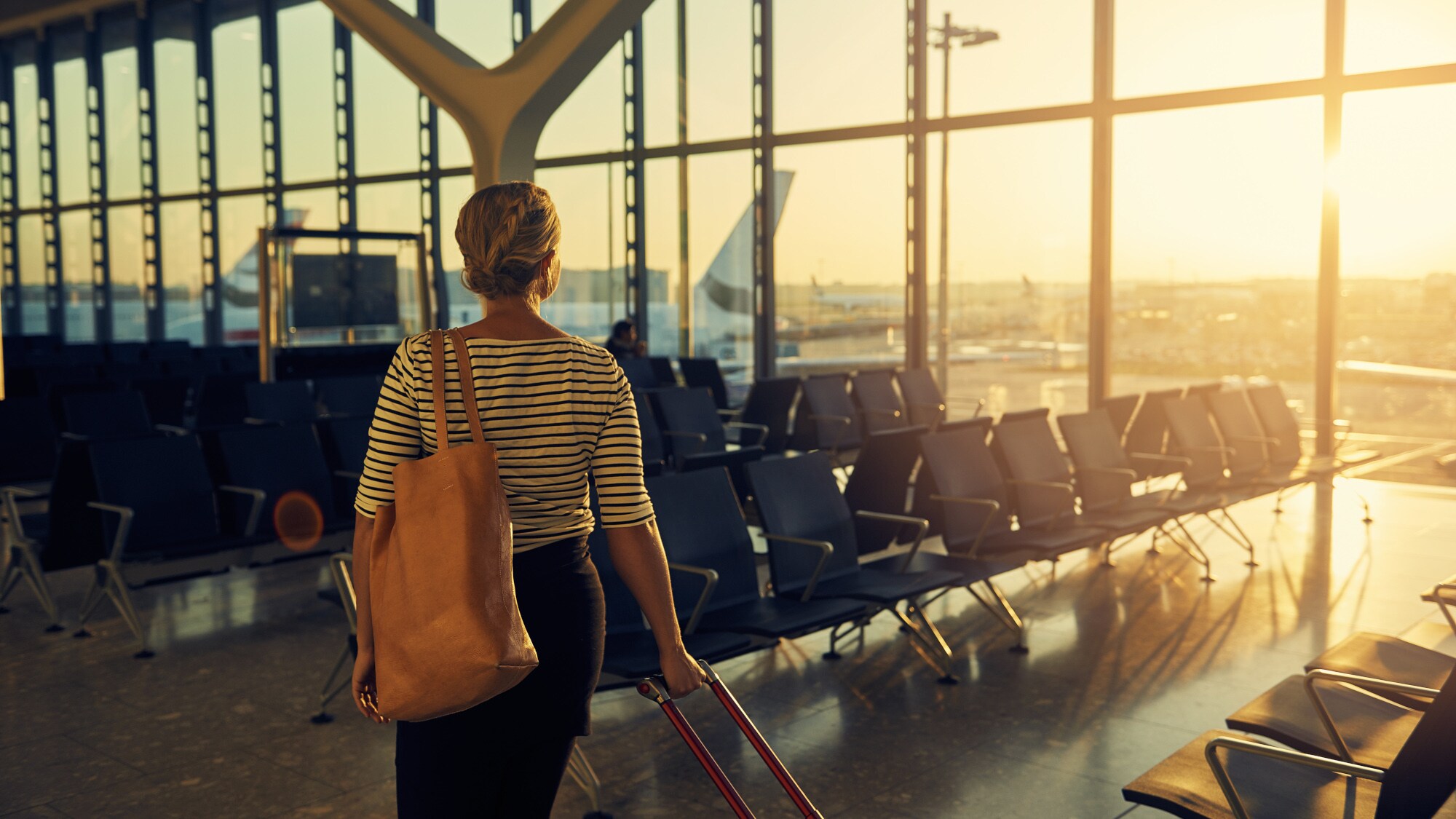 Rückenansicht einer Frau, die mit einen Koffer durch eine leere Wartehalle am Flughafen geht, im Hintergrund geht die Sonne unter.