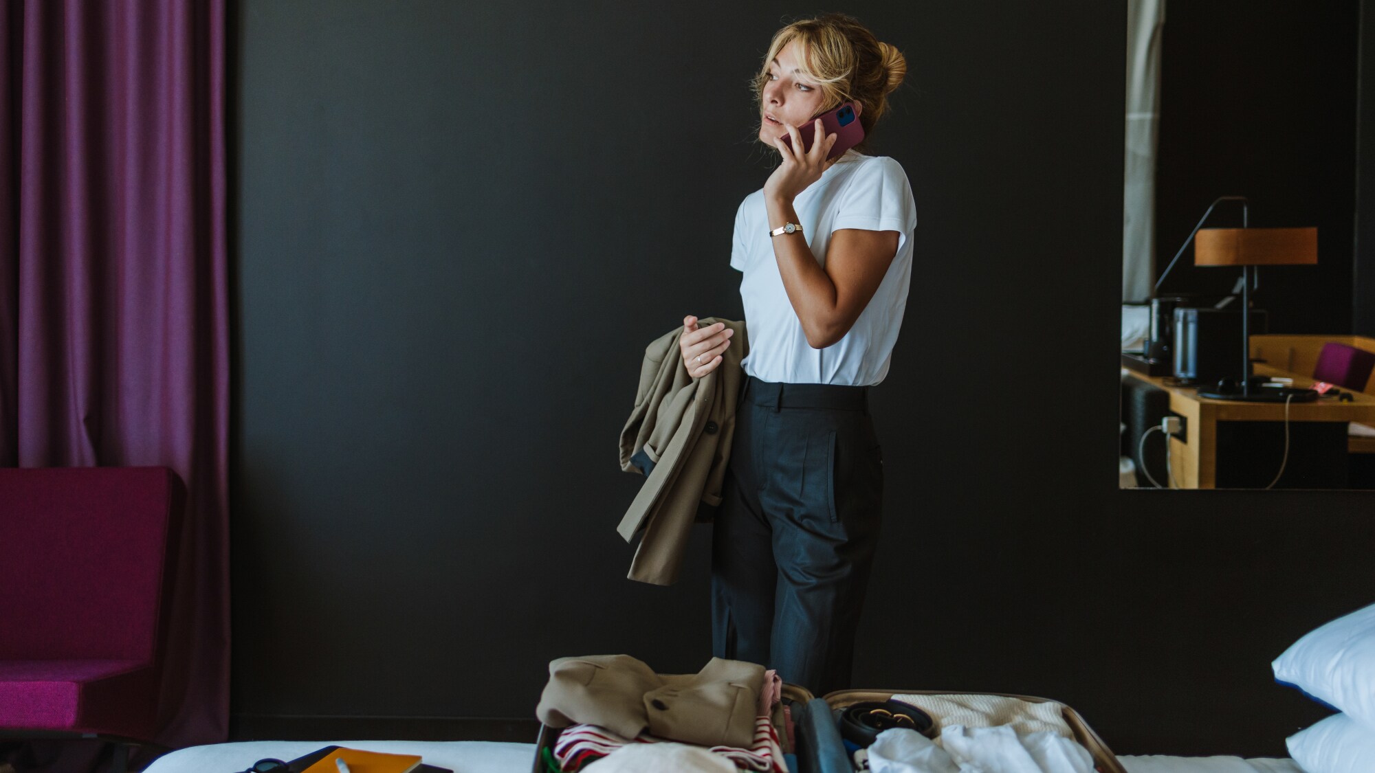 Eine Frau in einem Hotelzimmer, die ihr Smartphone ans Ohr hält und in der anderen Hand eine Jacke hält. Auf dem Bett liegt ein geöffneter Koffer mit verschiedenen Klamotten. Eine Frau in einem Hotelzimmer, die ihr Smartphone ans Ohr hält und in der anderen Hand eine Jacke hält. Auf dem Bett liegt ein geöffneter Koffer mit verschiedenen Klamotten.