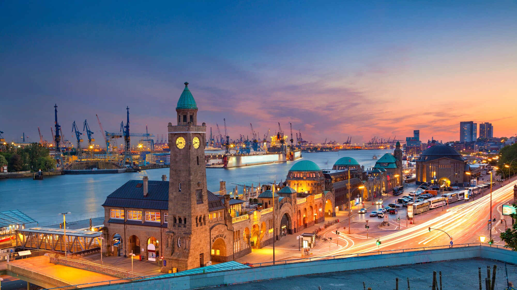 Der Wasserbahnhof Landungsbrücken im Hamburger Hafen bei Dämmerung mit Kränen des Containerhafens im Hintergrund. Der Wasserbahnhof Landungsbrücken im Hamburger Hafen bei Dämmerung mit Kränen des Containerhafens im Hintergrund.