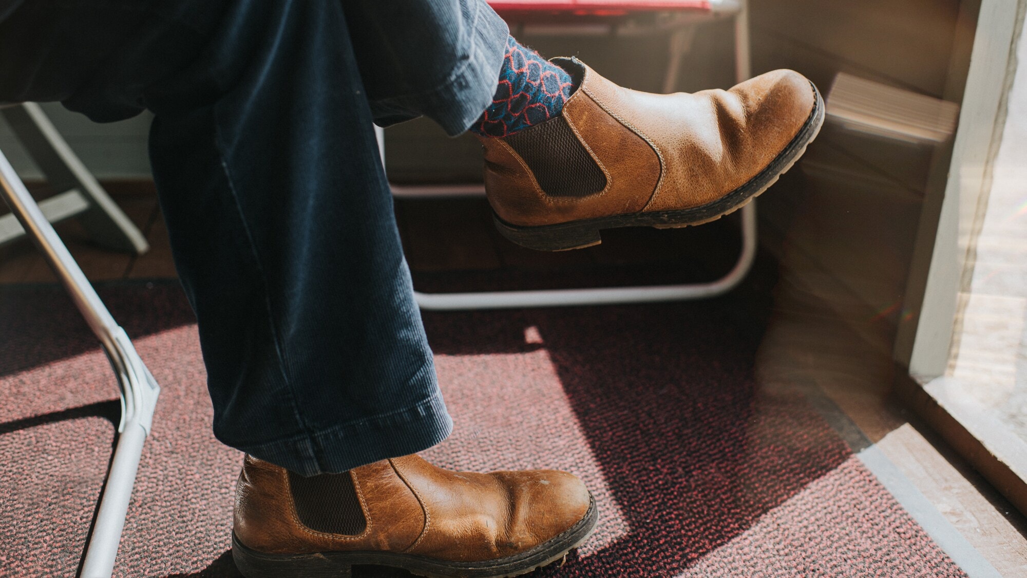 Mann sitzt mit gekreuzten Beinen und in hellbraunen Chelseaboots bei offener Tür in einem sonnendurchfluteten Raum. Mann sitzt mit gekreuzten Beinen und in hellbraunen Chelseaboots bei offener Tür in einem sonnendurchfluteten Raum.