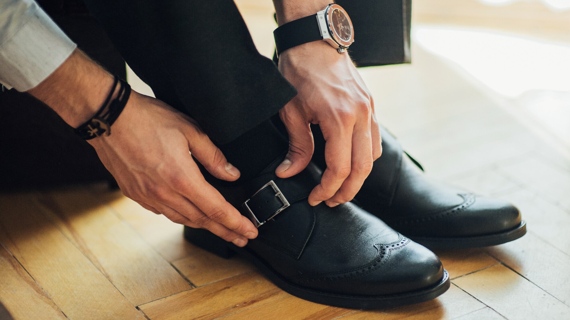 Männerhände schließen den seitlichen Schnallriemen eines schwarzen Monkstrap-Schuhs mit Lochmusternaht auf der Zehenkappe. Männerhände schließen den seitlichen Schnallriemen eines schwarzen Monkstrap-Schuhs mit Lochmusternaht auf der Zehenkappe.
