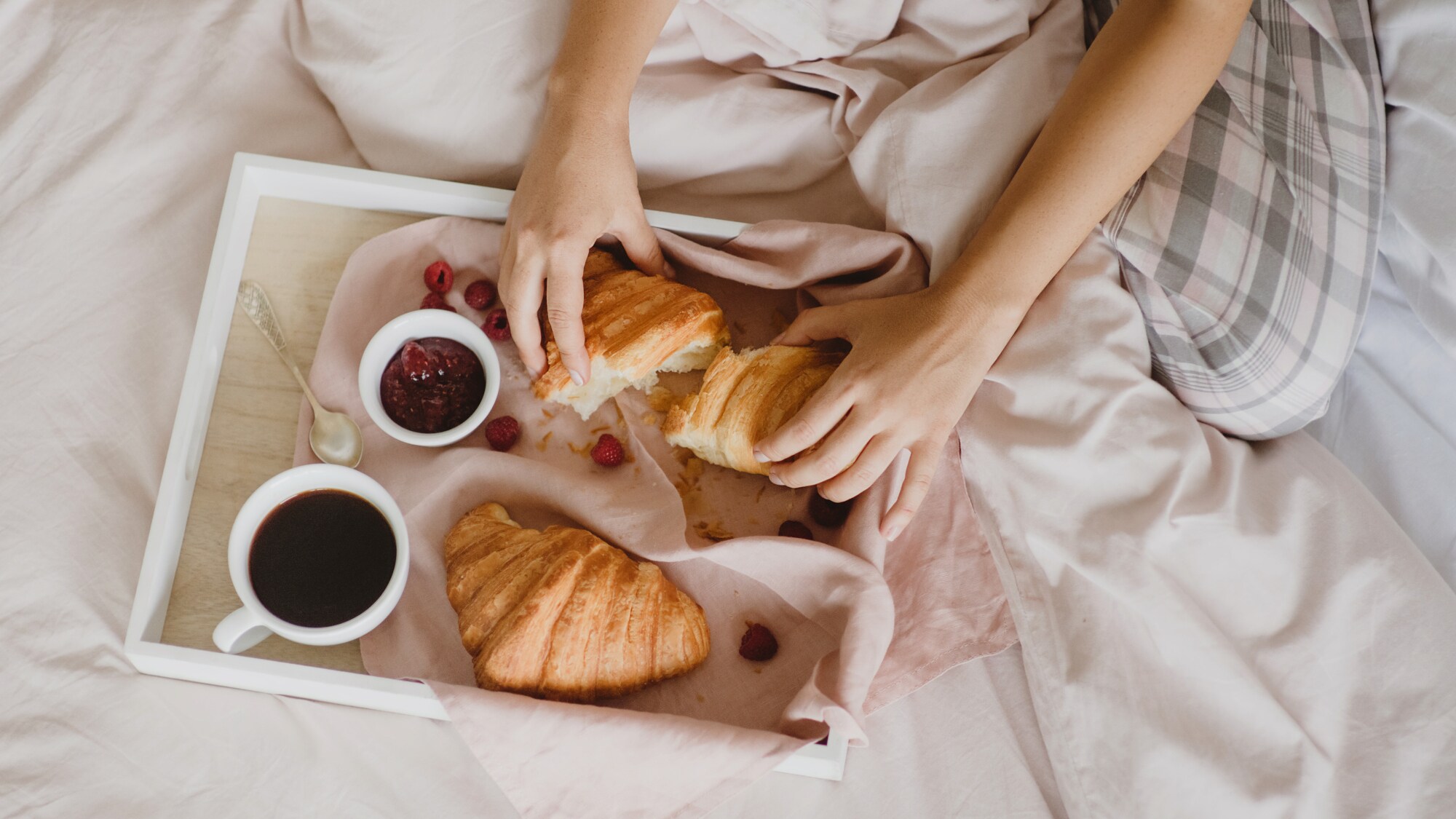 Auf einem Bett liegt ein Tablett mit Croissants, einer Tasse Kaffee, ein Schälchen Marmelade, einem Löffel und Früchten. Dahinter sitzt eine unkenntliche Person, die eines der Croissants auseinanderreißt.
