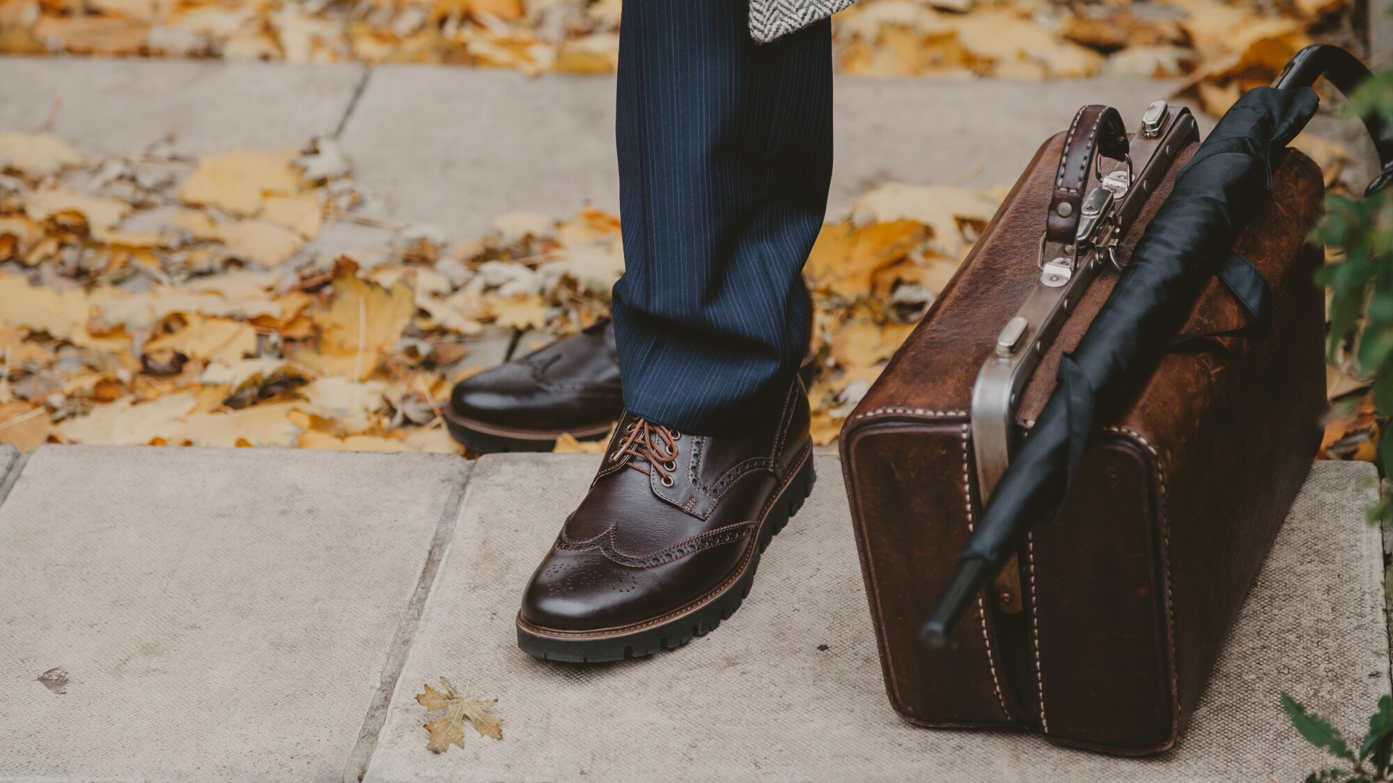 Männliche Beine in gestreifter Hose und braunen Budapesterschuhen auf betoniertem Untergrund mit Herbstblättern, daneben eine alte Doktortasche mit darauf liegendem Stockschirm. Männliche Beine in gestreifter Hose und braunen Budapesterschuhen auf betoniertem Untergrund mit Herbstblättern, daneben eine alte Doktortasche mit darauf liegendem Stockschirm.