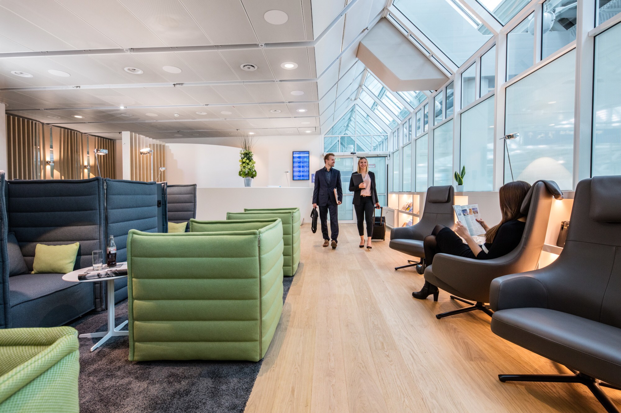 Mehrere Personen in einer modernen Flughafen-Lounge mit Sesseln vor einem Panoramafenster.