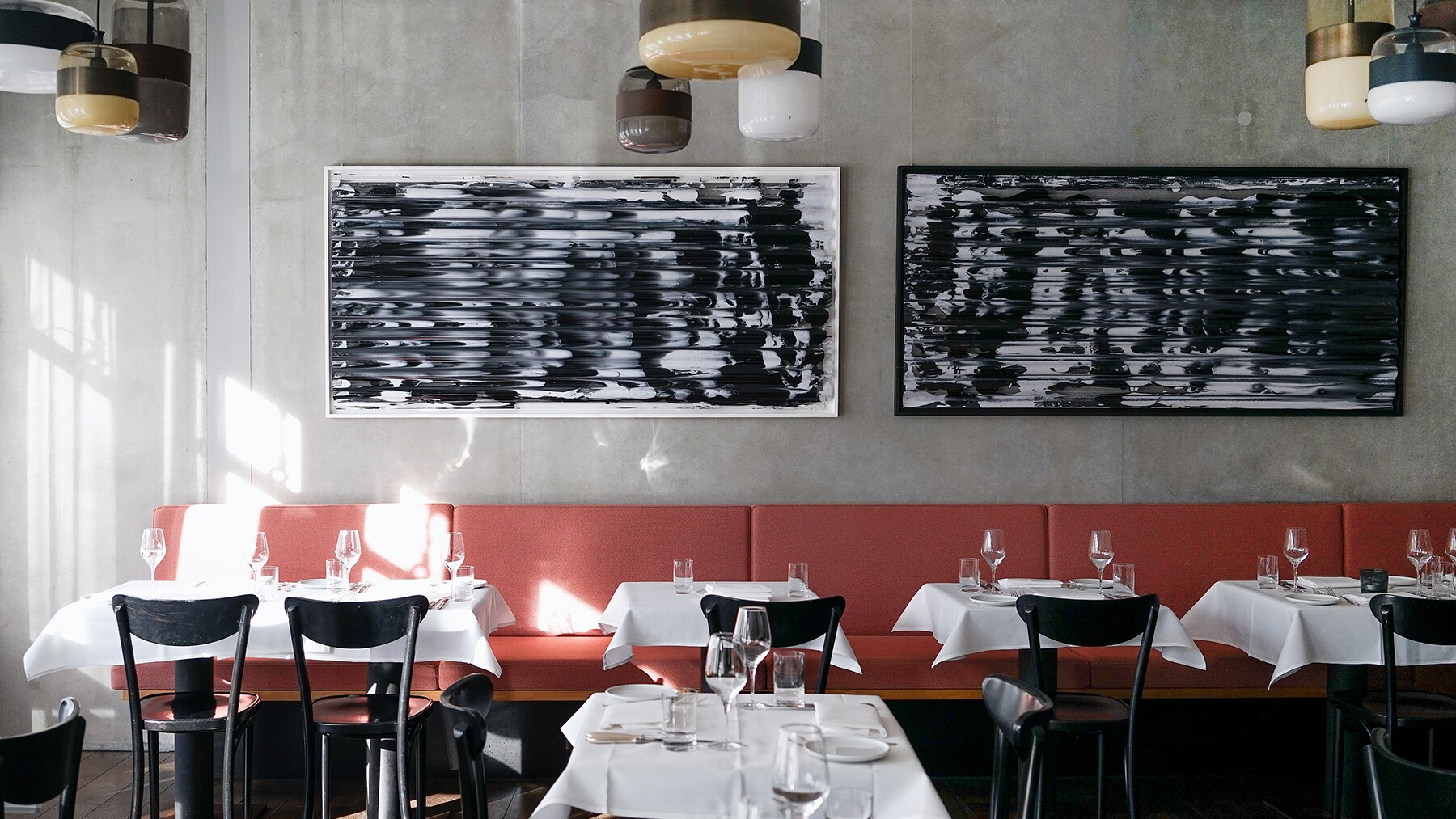 Ein lichtdurchfluteter Raum mit mehreren eingedeckten Tischen, weißen Tischdecken, schwarzen Stühlen, einer rot-braunen durchgängigen Sitzbank und zwei großen Bildern mit abstrakten Schwarz-Weiß-Motiven.
