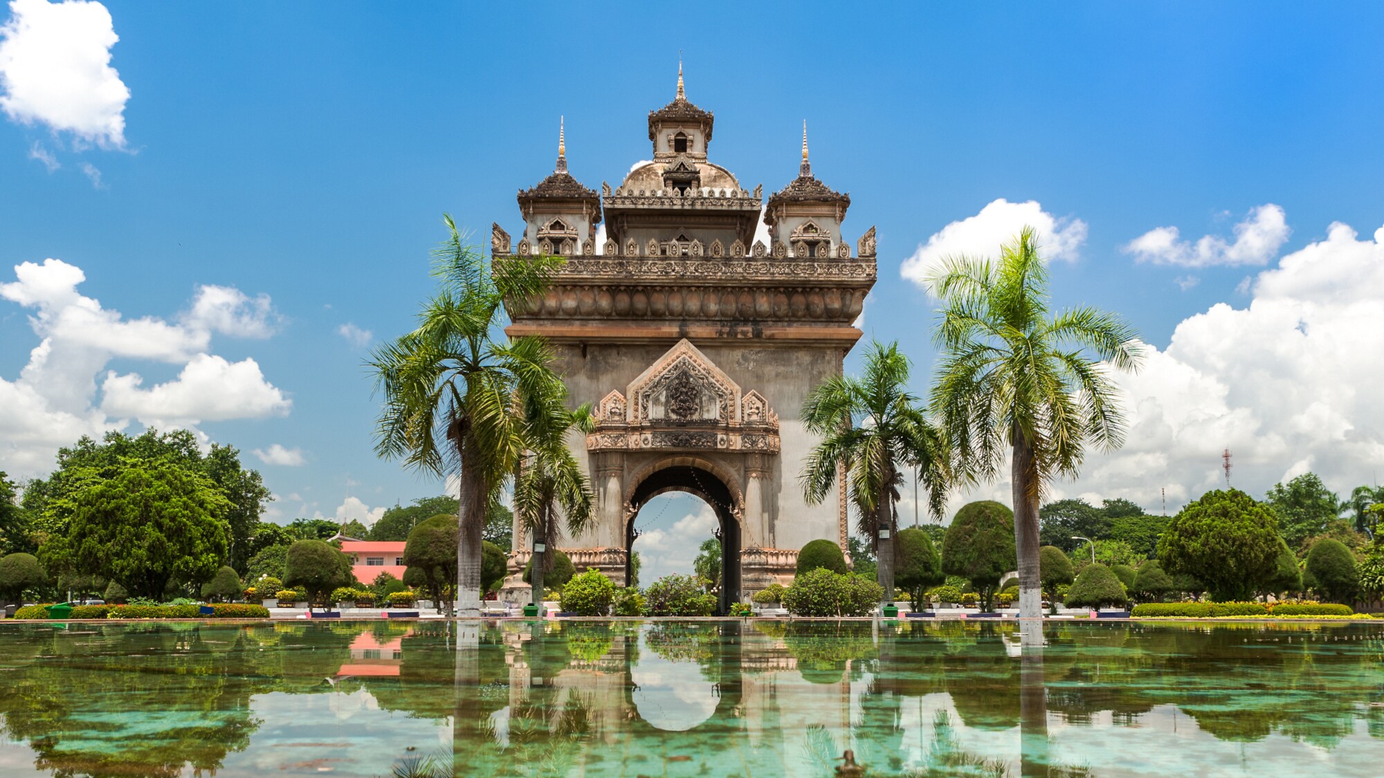 Das Patuxai-Denkmal in Vientiane, Laos. Davor Wasser mit Spiegelungen, blauer Himmel mit Wolken.