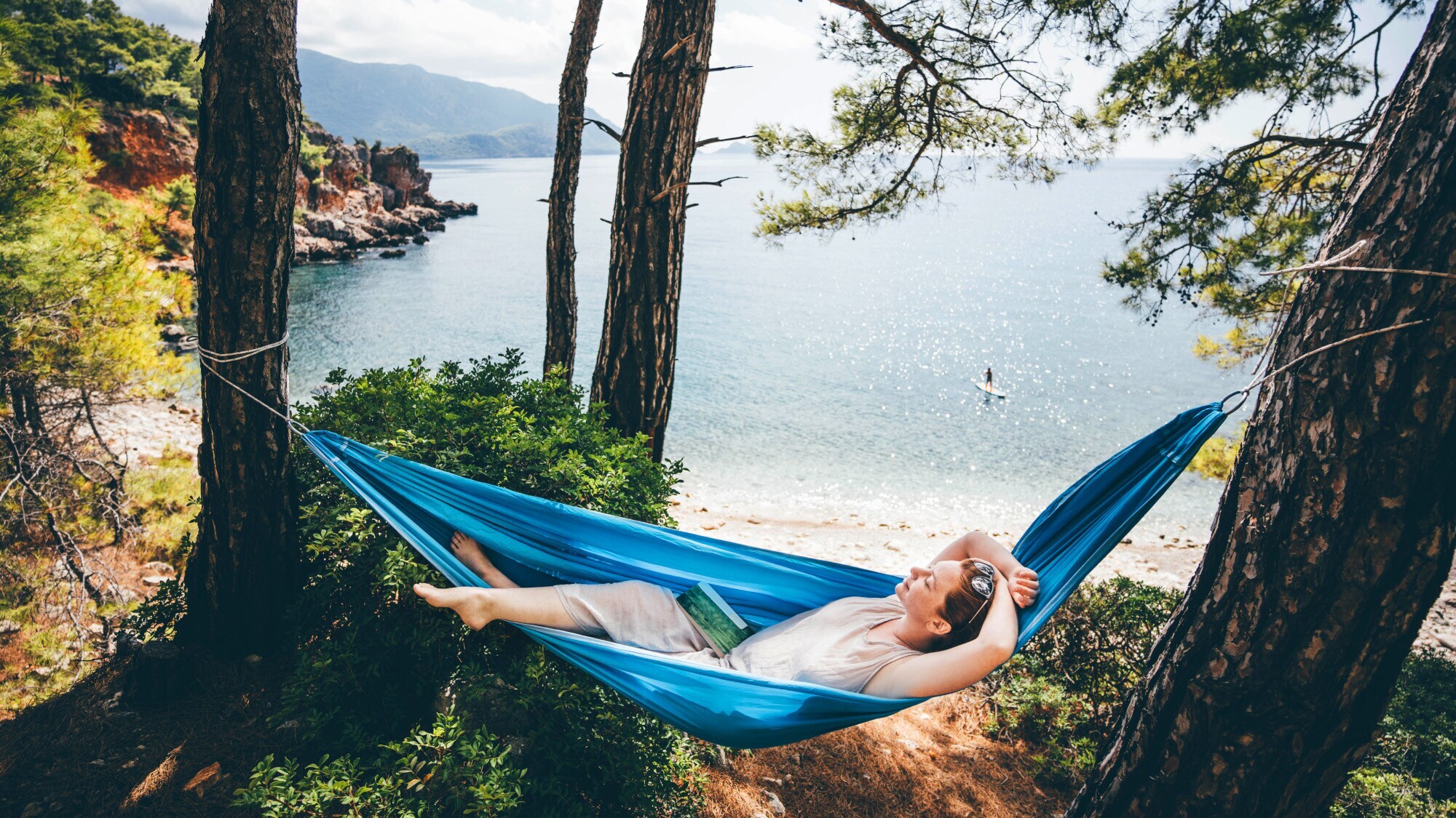 Eine Frau entspannt in einer blauen Hängematte zwischen Pinien an einer Meeresküste.