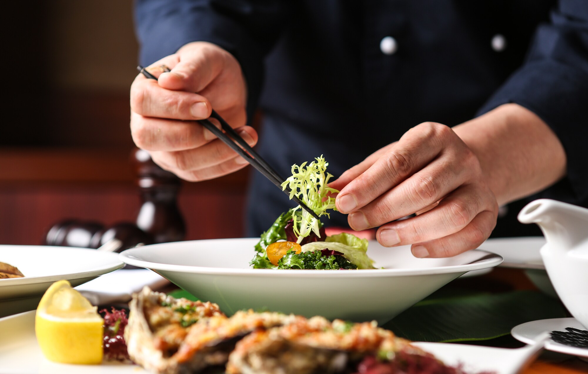 Zwei Hände bereiten auf einem weißen Teller eine Speise mit Essstäbchen zu. Zwei Hände bereiten auf einem weißen Teller eine Speise mit Essstäbchen zu.