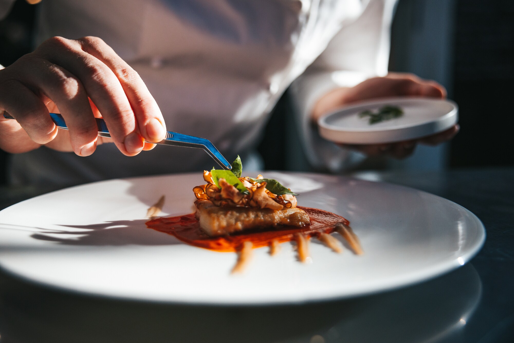 Ein Gourmetgericht wird von einer Person in Kochjacke mit einer Pinzette auf einem weißen Teller angerichtet.