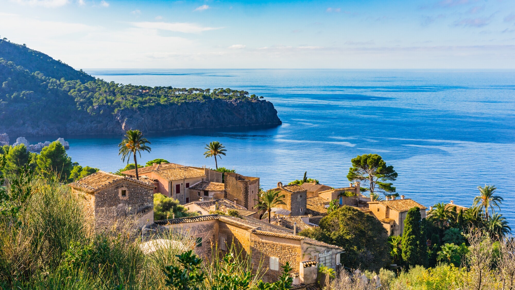 Ein altes Dorf an der Küste Mallorcas. Im Hintergrund eine grün überwachsene Landzunge, die in das Meer hineinragt.
