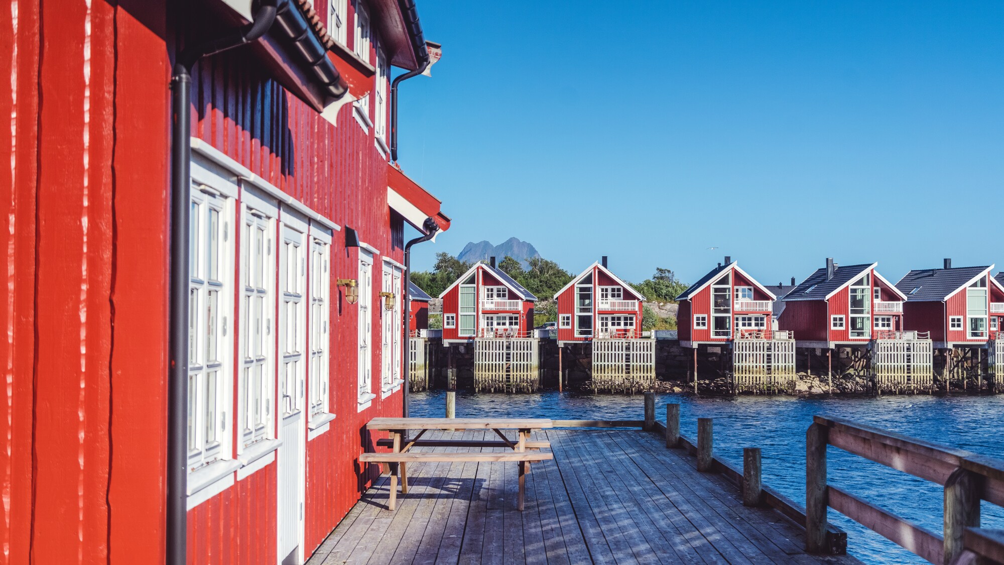 Eine Reihe roter Fischerhütten auf den Lofoten in Norwegen. Im Vordergrund ist eine Hütte mit Terrasse, Sitzbank und Tisch im Fokus. Eine Reihe roter Fischerhütten auf den Lofoten in Norwegen. Im Vordergrund ist eine Hütte mit Terrasse, Sitzbank und Tisch im Fokus.