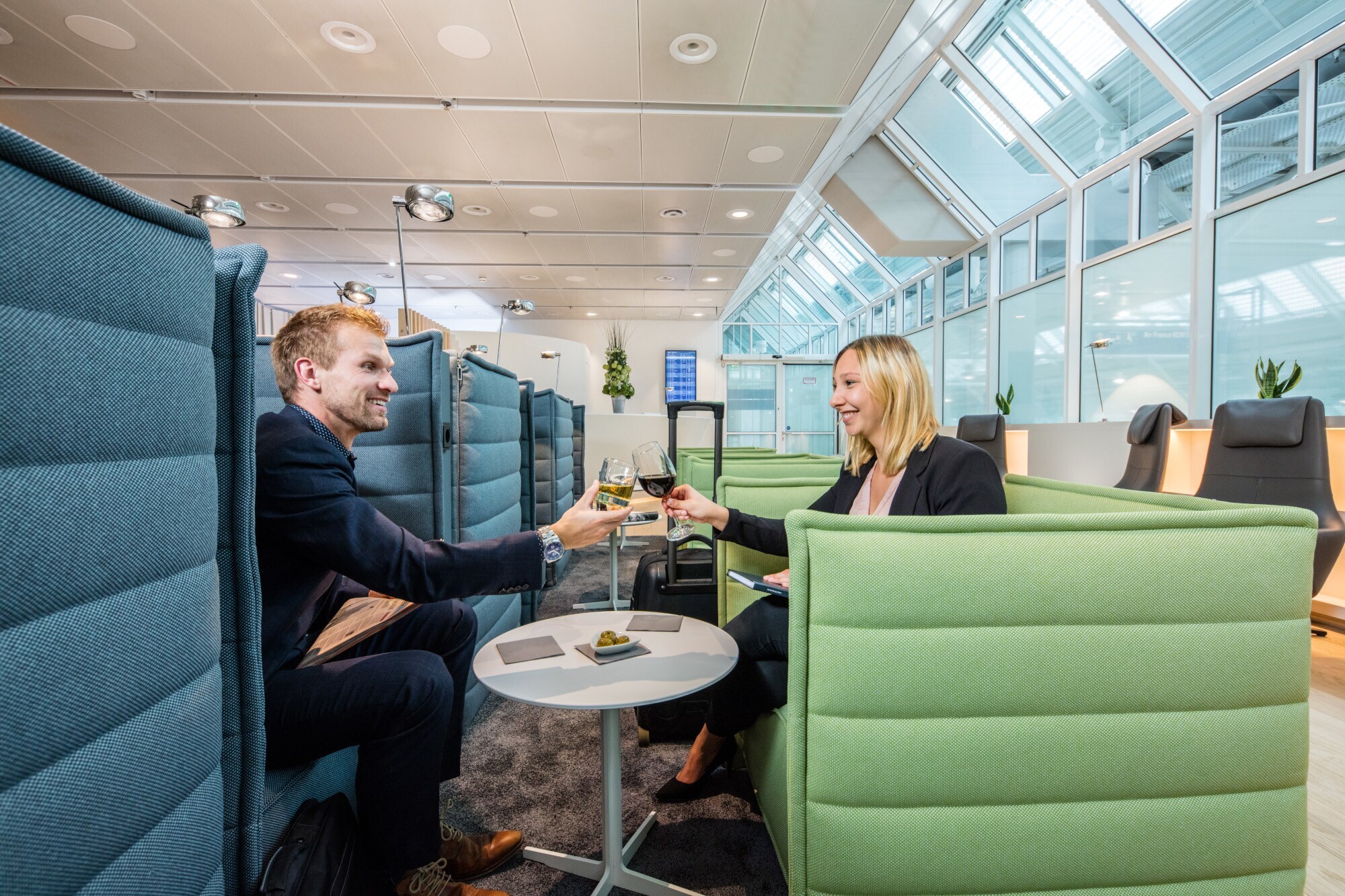 Ein Mann und eine Frau sitzen sich gegenüber auf Sesseln in einer Flughafenlounge und stoßen mit Weingläsern an.