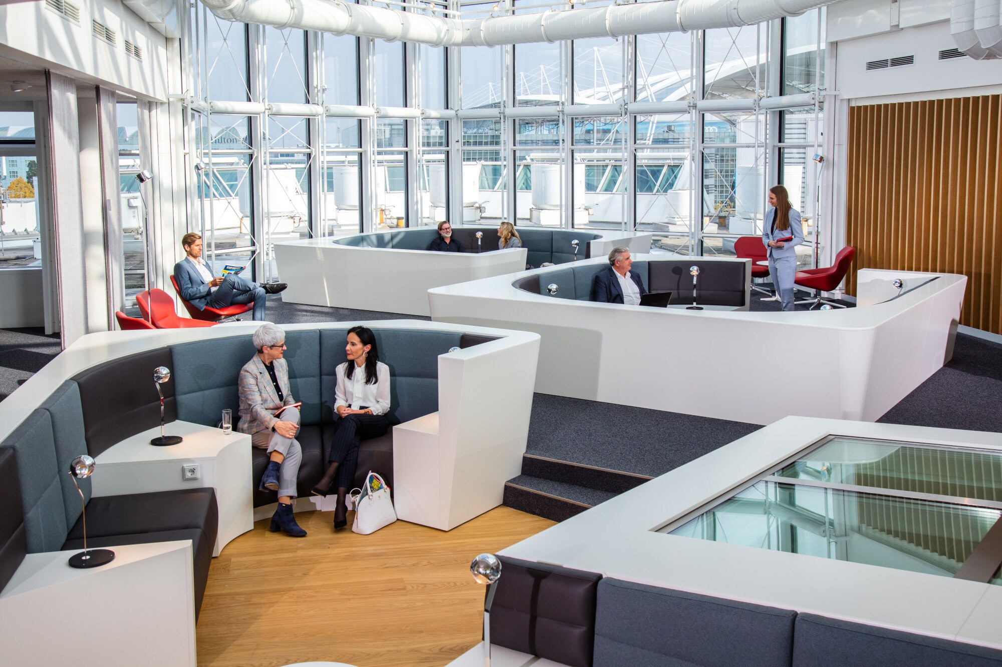 Mehrere Personen sitzen in einer modernen Sitzlandschaft einer Flughafenlounge mit Panoramafenstern.