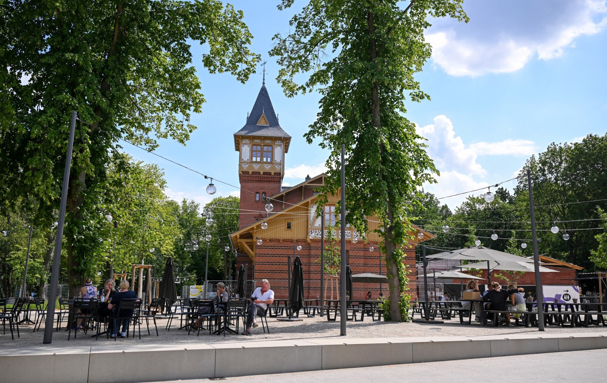 Personen in einem Biergarten vor einem Backsteinhaus mit Turm.