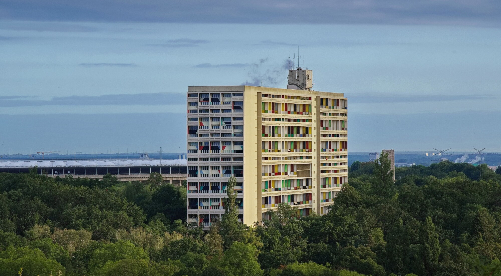 Luftaufnahme eines von Wald umgebenen Hochhauses im Corbusier-Stil mit gemusterter Fassade vor einem Stadion. Luftaufnahme eines von Wald umgebenen Hochhauses im Corbusier-Stil mit gemusterter Fassade vor einem Stadion.