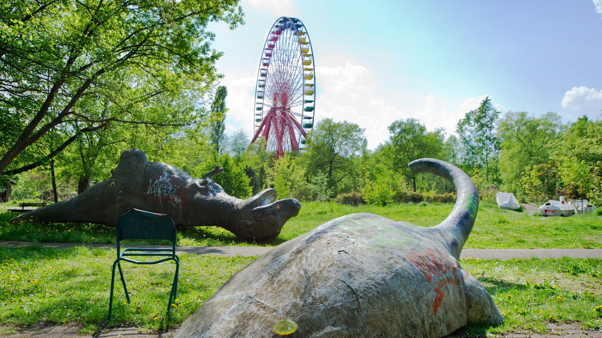 Zwei umgekippte Dinosaurier-Skulpturen auf einer Wiese vor einem Riesenrad in einem Freizeitpark. Zwei umgekippte Dinosaurier-Skulpturen auf einer Wiese vor einem Riesenrad in einem Freizeitpark.