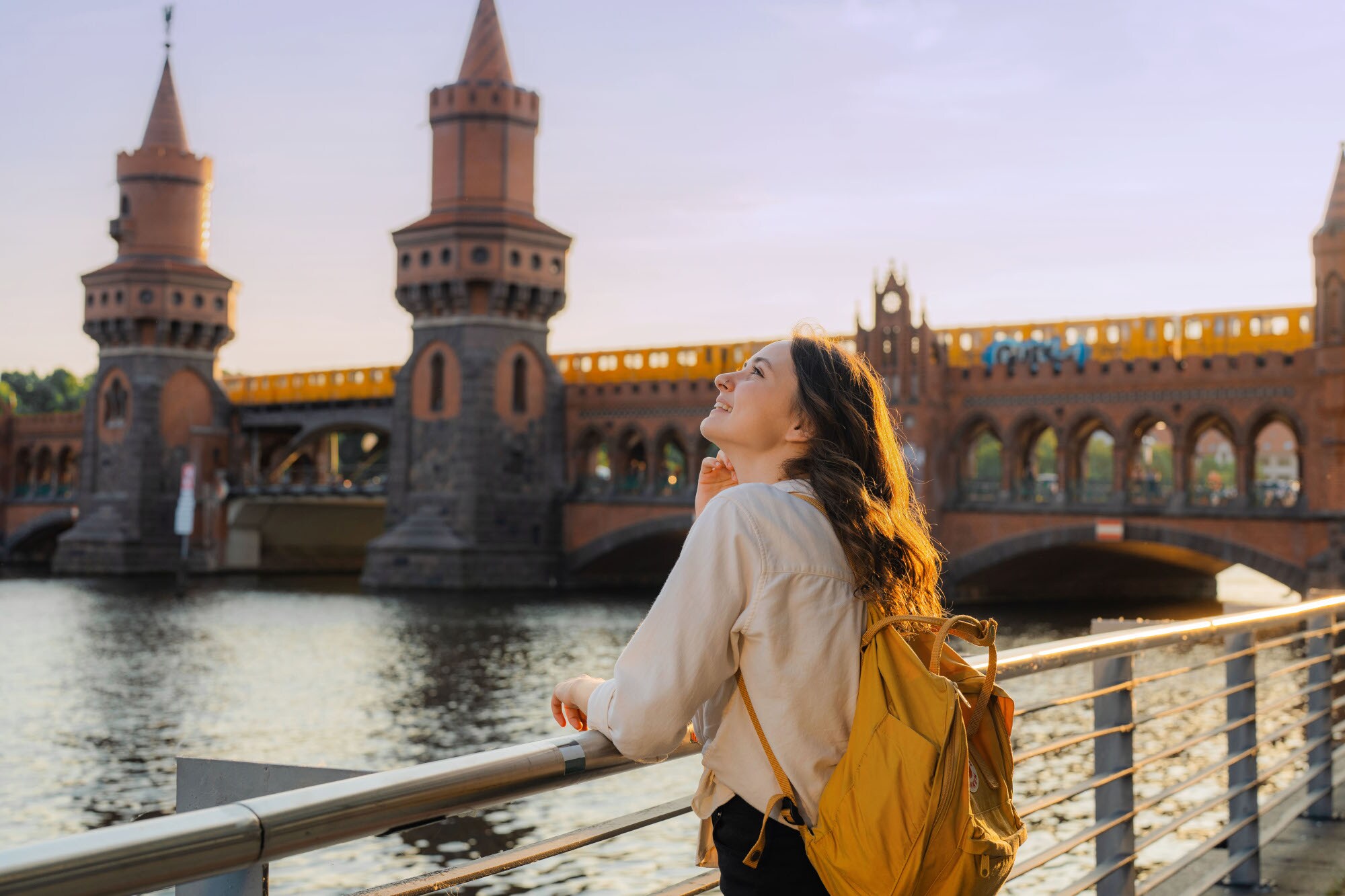Eine junge Frau mit Rucksack steht am Wasser vor einer Brücke mit Türmen, auf der eine gelbe Straßenbahn fährt. Eine junge Frau mit Rucksack steht am Wasser vor einer Brücke mit Türmen, auf der eine gelbe Straßenbahn fährt.