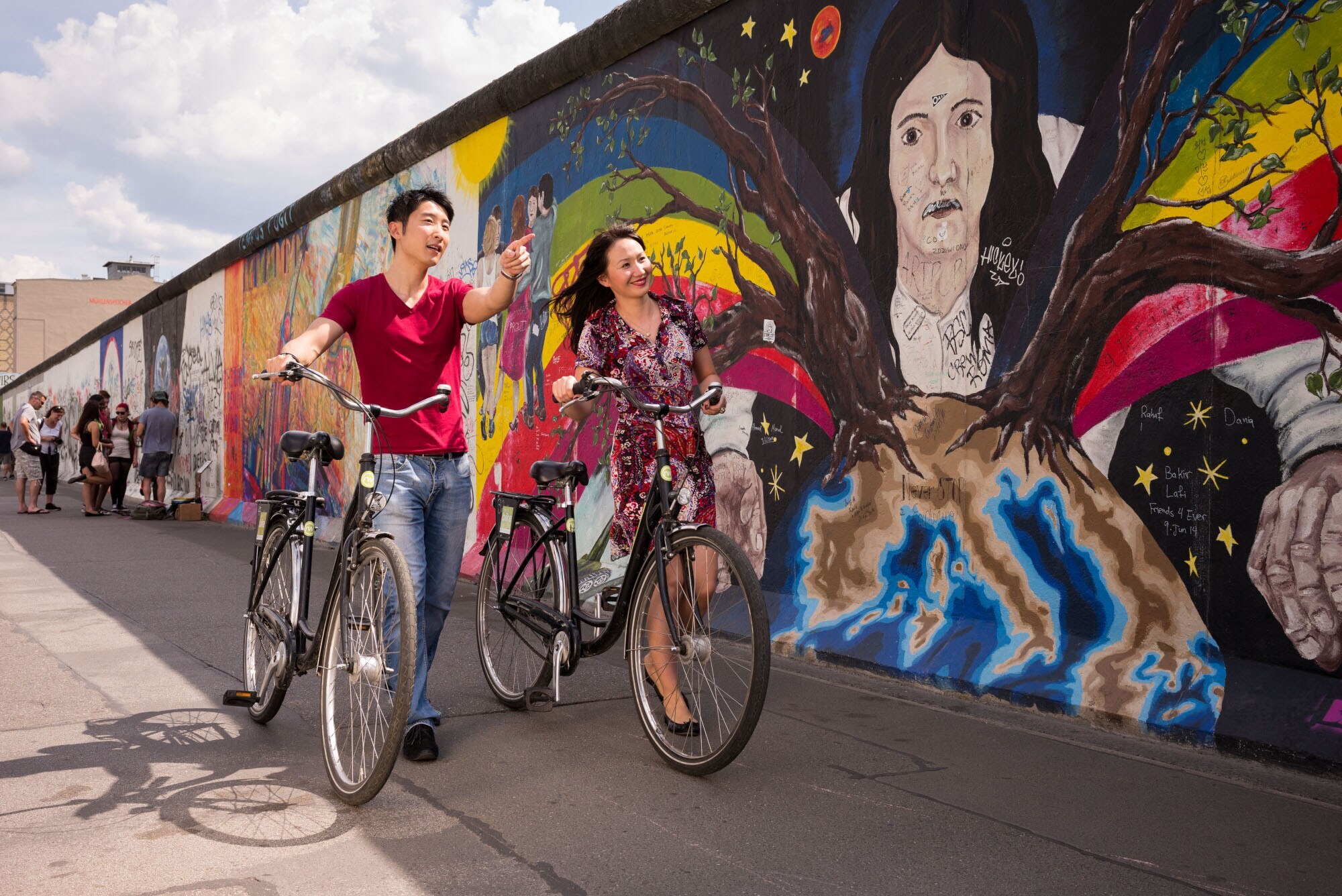 Ein Mann und eine Frau schieben jeweils ein Fahrrad an einem Kunstwerk an der Berliner Mauer entlang. Ein Mann und eine Frau schieben jeweils ein Fahrrad an einem Kunstwerk an der Berliner Mauer entlang.