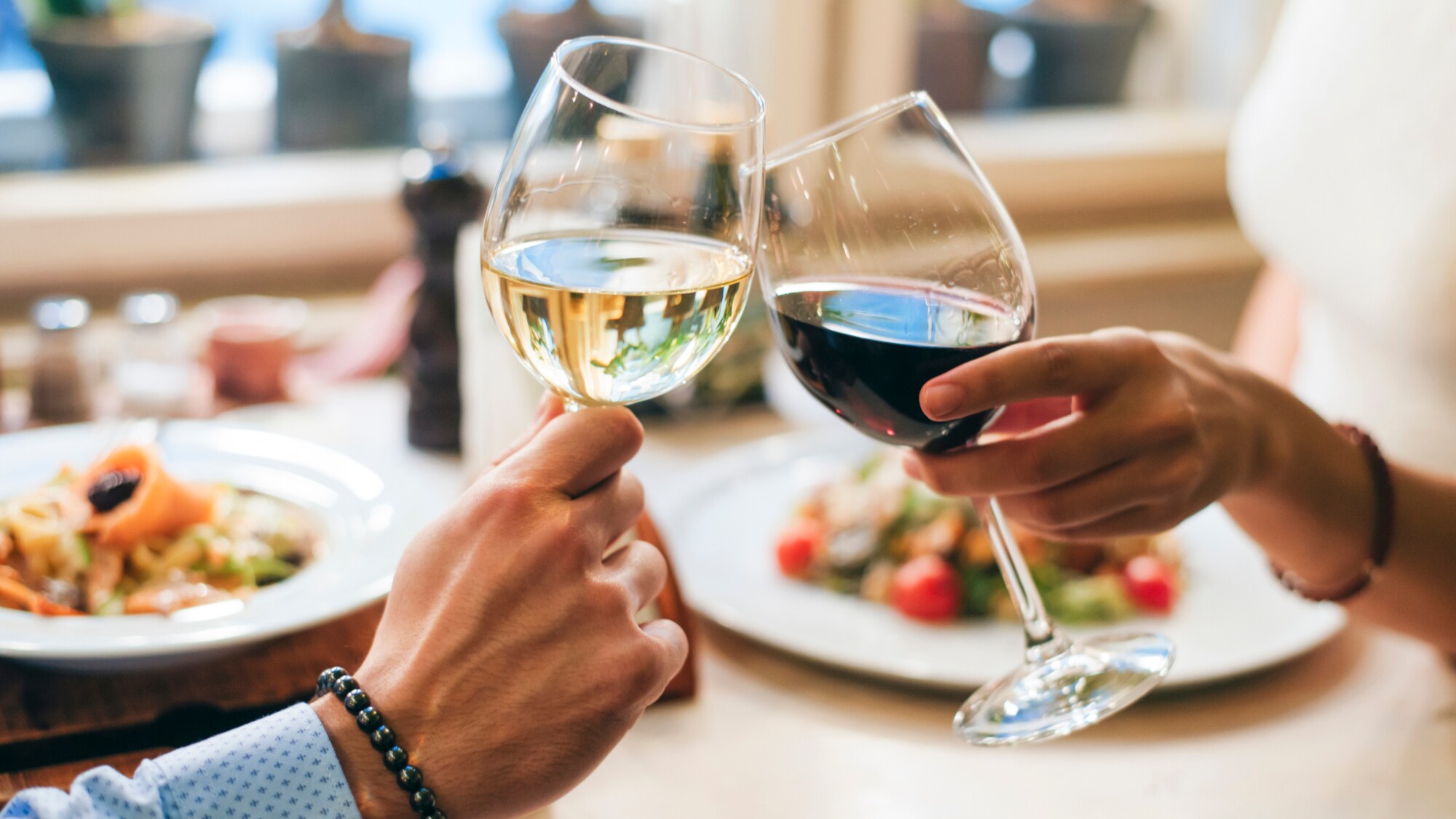 Zwei Personen, die jeweils ein Glas Wein halten und anstoßen. Auf dem Tisch vor ihnen stehen Teller mit Speisen.