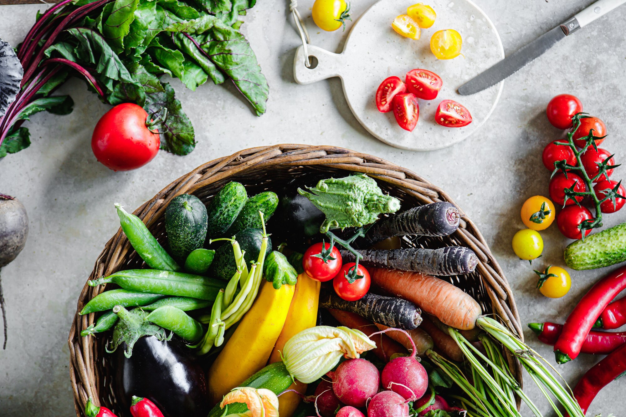 Verschiedene Gemüsesorten liegen auf einem hellen Tisch und in einem großen Korb, der auf dem Tisch steht.