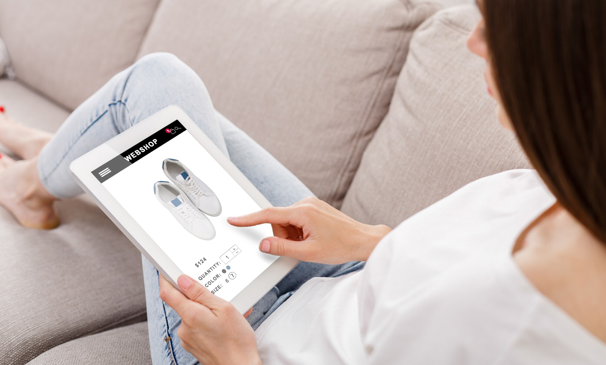 Eine Frau liegt mit einem Tablet auf der Couch und betrachtet weiße Sneaker in einem Onlineshop.