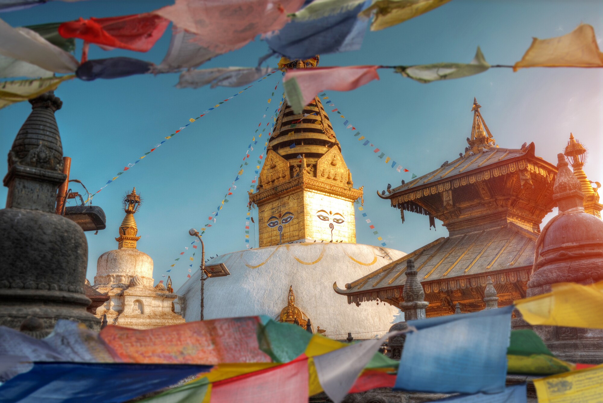 Goldener Turm einer buddhistische Tempelanlage mit aufgemalten Gesichtern neben einer Pagode, im Vordergrund wehende, bunte Gebetsflaggen. Goldener Turm einer buddhistische Tempelanlage mit aufgemalten Gesichtern neben einer Pagode, im Vordergrund wehende, bunte Gebetsflaggen.