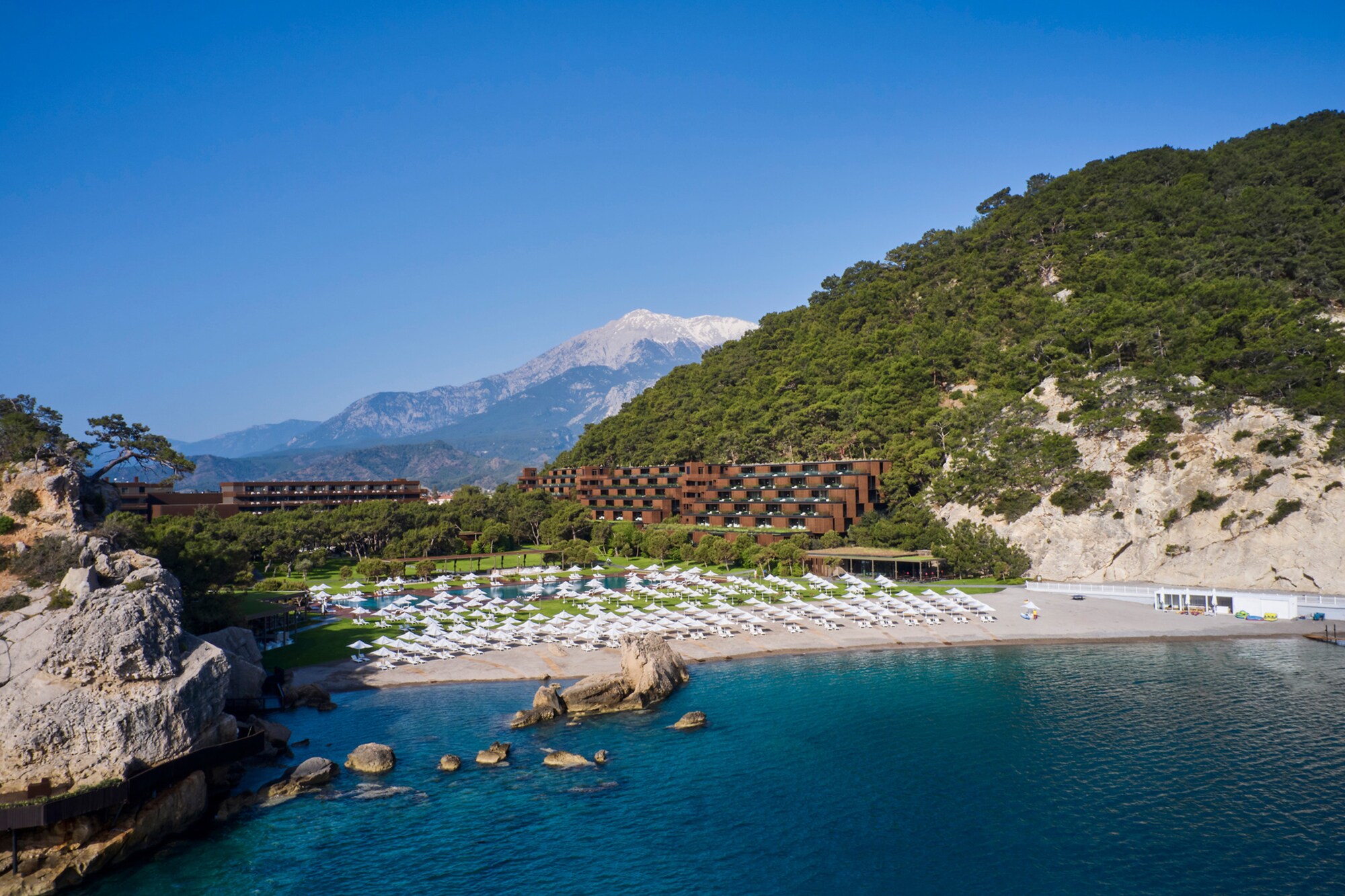 Ein Hotelresort in einer mediterranen Bucht mit Sandstrand, im Hintergrund eine Berglandschaft.