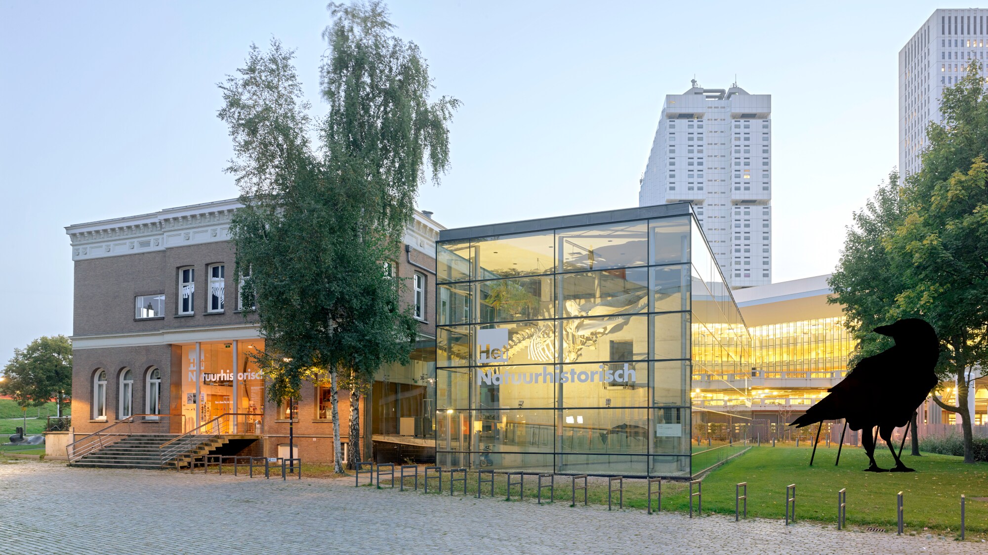 Ein beleuchteter Gebäudekomplex eines Naturkundemuseums mit Glaspavillon in dem ein Dinosaurierskelett hängt, daneben eine Grünfläche mit einer Skulptur in Vogelform. Ein beleuchteter Gebäudekomplex eines Naturkundemuseums mit Glaspavillon in dem ein Dinosaurierskelett hängt, daneben eine Grünfläche mit einer Skulptur in Vogelform.