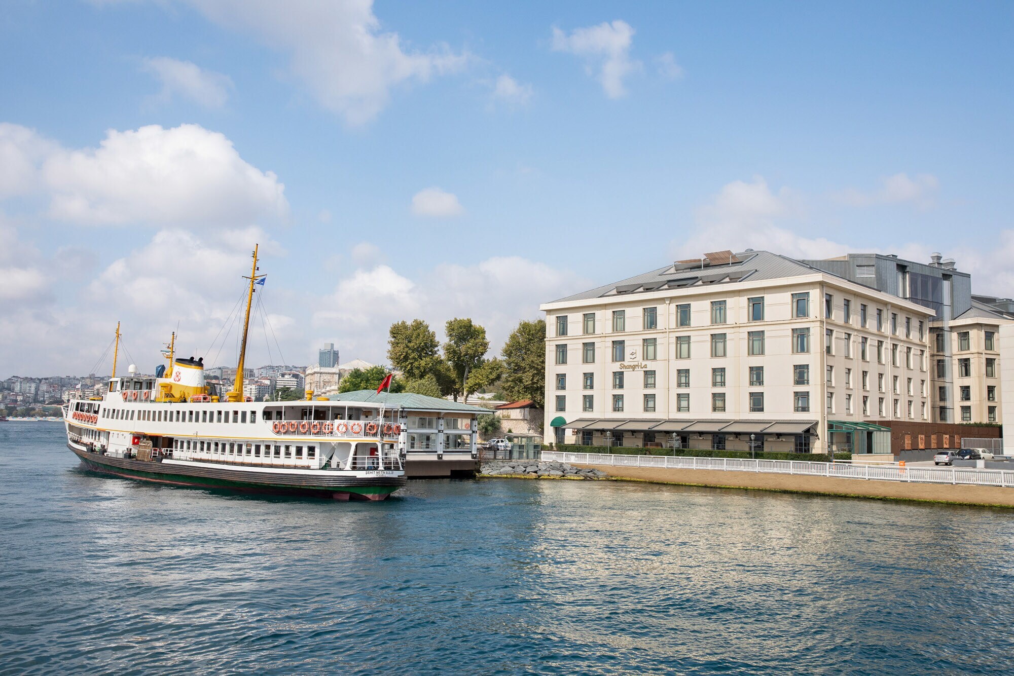 Gebäude mit neoklassizistischer Fassade am Ufer eines Gewässers mit Schiff im Vordergrund, im Hintergrund die Skyline von Istanbul.