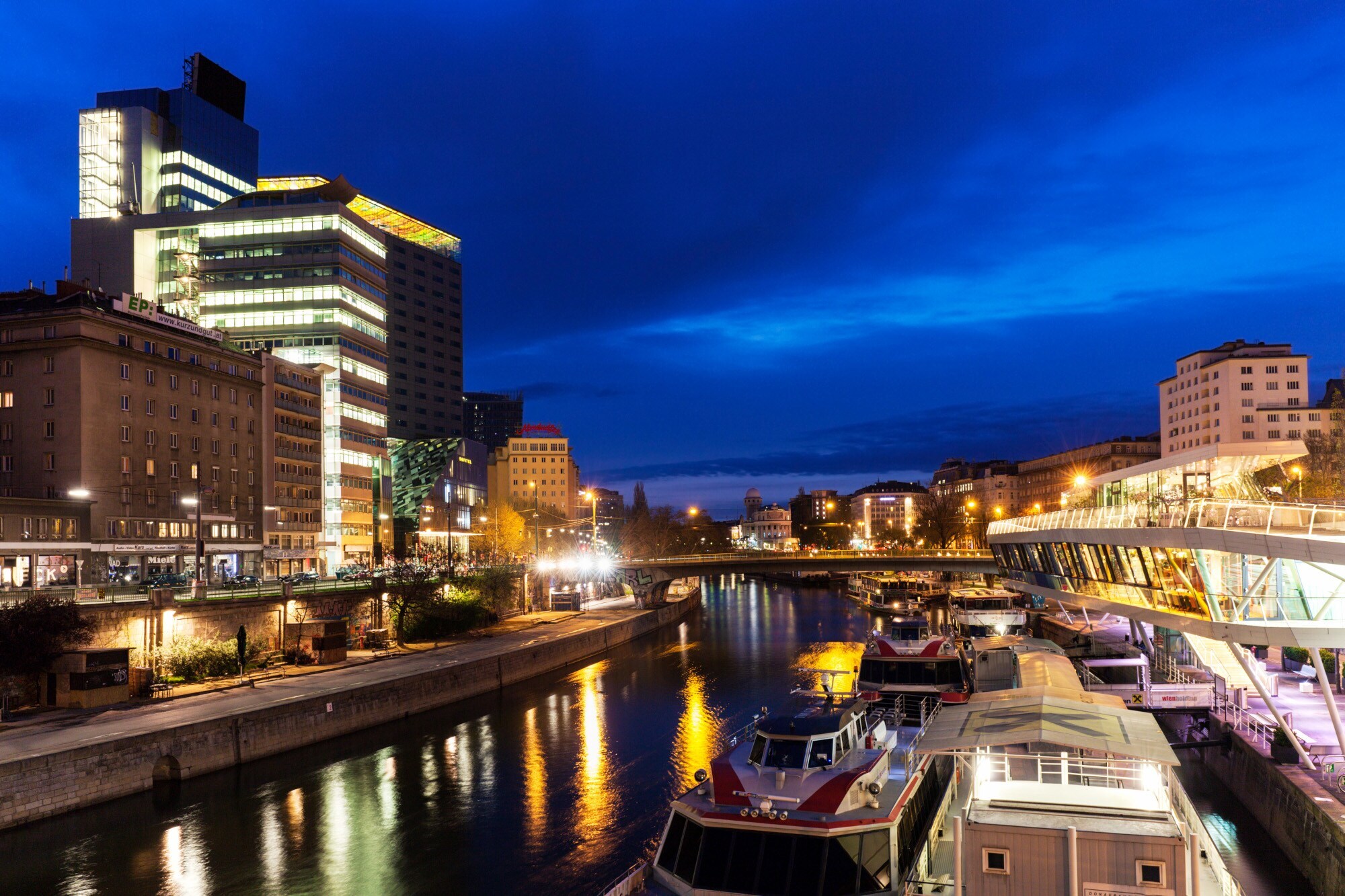 Moderner Hotelkomplex am Donaukanal mit Barkassen in Wien bei Nacht.