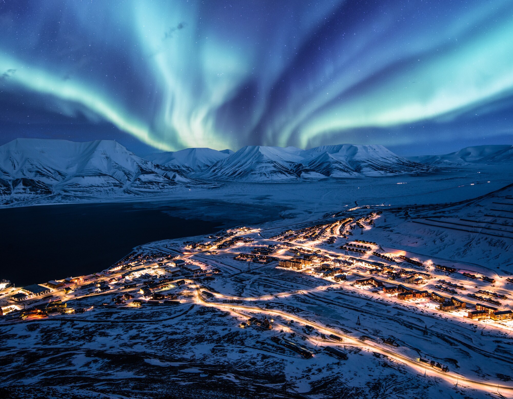 Beleuchtete Ortschaft in schneebedeckter Fjordlandschaft unter Polarlichtern bei Nacht. Beleuchtete Ortschaft in schneebedeckter Fjordlandschaft unter Polarlichtern bei Nacht.