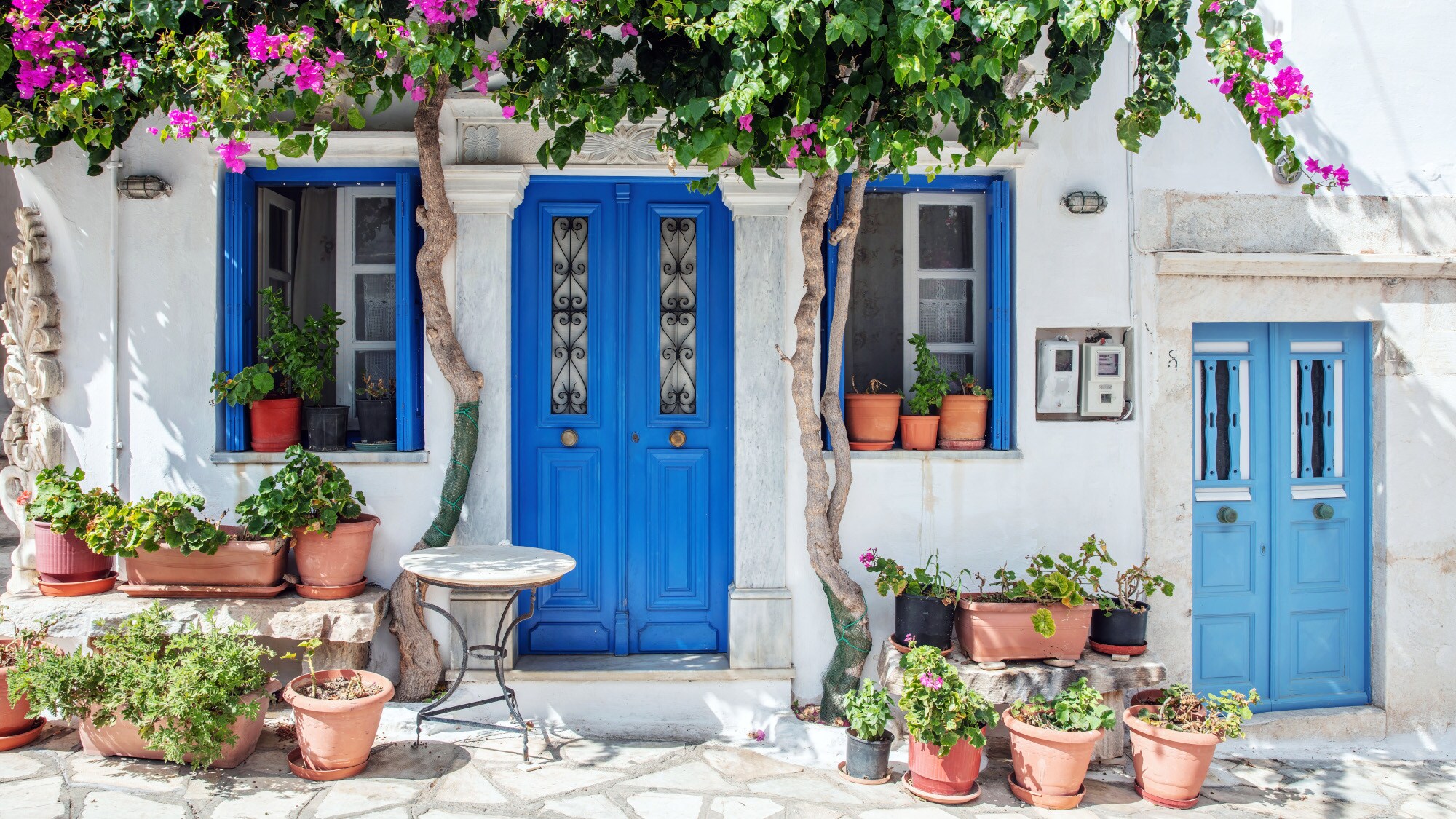 Weißes Haus im kykladischen Stil mit blauen Fenstern und Türen, davor Pflanzentöpfe und blühender Bougainvillea. Weißes Haus im kykladischen Stil mit blauen Fenstern und Türen, davor Pflanzentöpfe und blühender Bougainvillea.