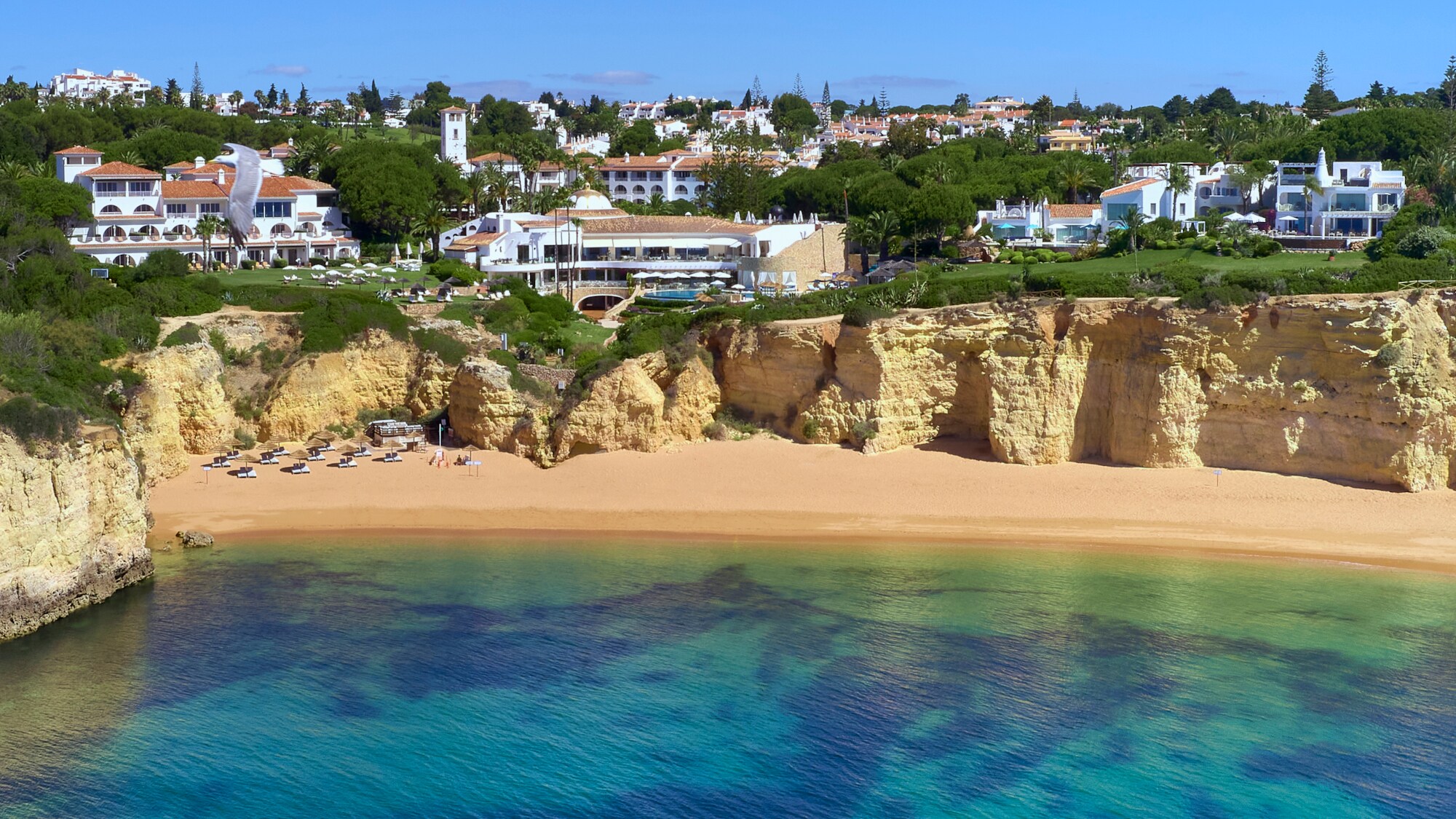 Luftaufnahme der Hotelanlage des Vila Vita Parc, das direkt an einer Klippe liegt. Darunter befindet sich ein Sandstrand und türkisblaues Meer.