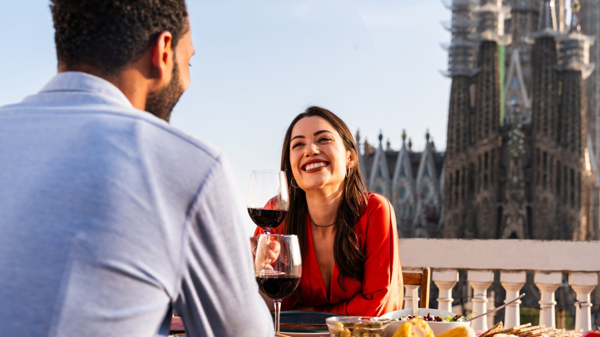 Eine Frau sitzt gegenüber von einem Mann an einem Tisch und hält lächelnd ein Glas Rotwein in der Hnad. Im Hintergrund die Sagrada Familia von Barcelona. Eine Frau sitzt gegenüber von einem Mann an einem Tisch und hält lächelnd ein Glas Rotwein in der Hnad. Im Hintergrund die Sagrada Familia von Barcelona.