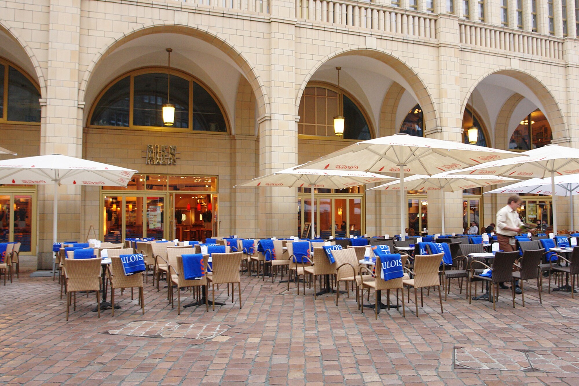 Außengastronomie auf einem Platz vor einem Gebäudekomplex mit Rundbögen.