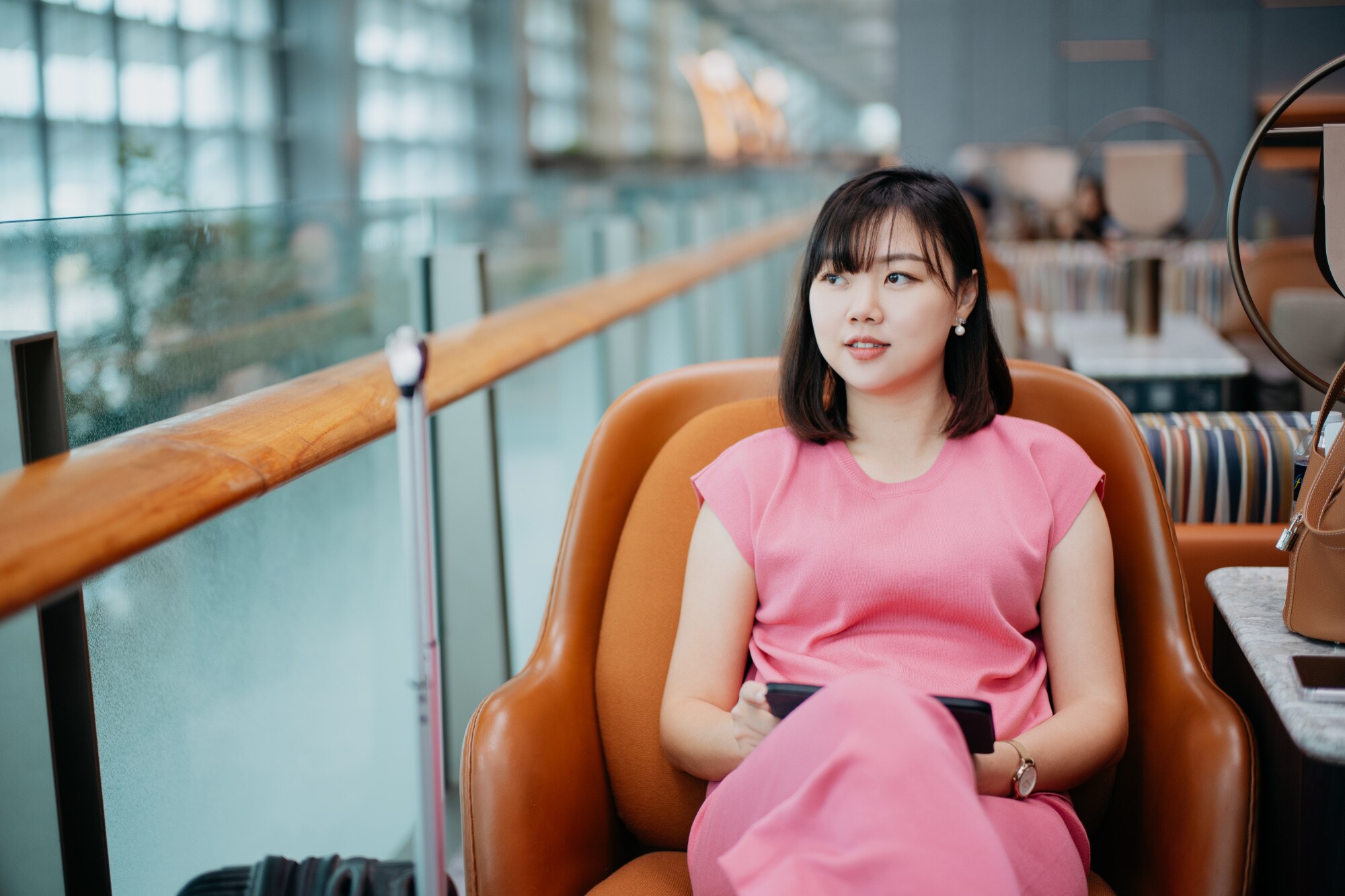 Eine asiatische Frau in einem pinken Kleid sitzt in einem braunen Ledersessel auf einer Galerie einer Flughafenlounge. Eine asiatische Frau in einem pinken Kleid sitzt in einem braunen Ledersessel auf einer Galerie einer Flughafenlounge.