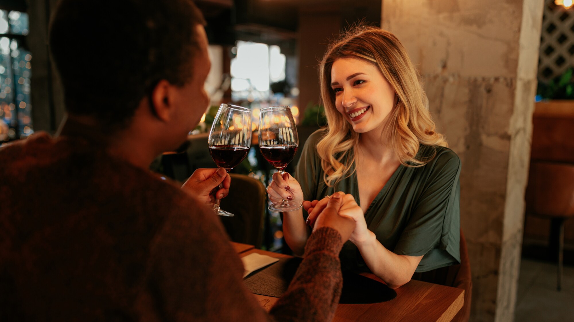 Eine lächelnde Frau sitzt einem Mann an einem Restauranttisch gegenüber und hält seine Hand. In der anderen Hand halten sie je ein Glas Rotwein.