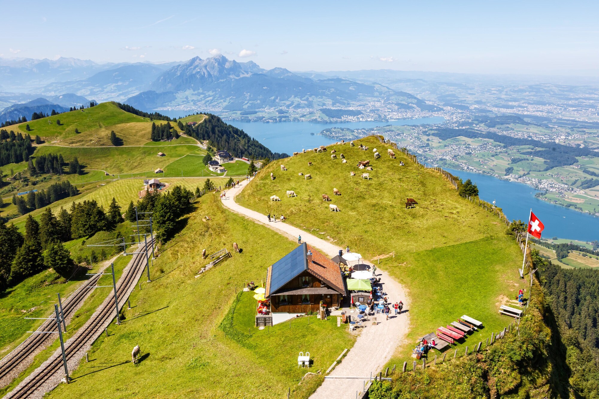 Aufsicht eines grünen Berggipfels mit Almhütte und Kühen oberhalb einer Seenlandschaft.