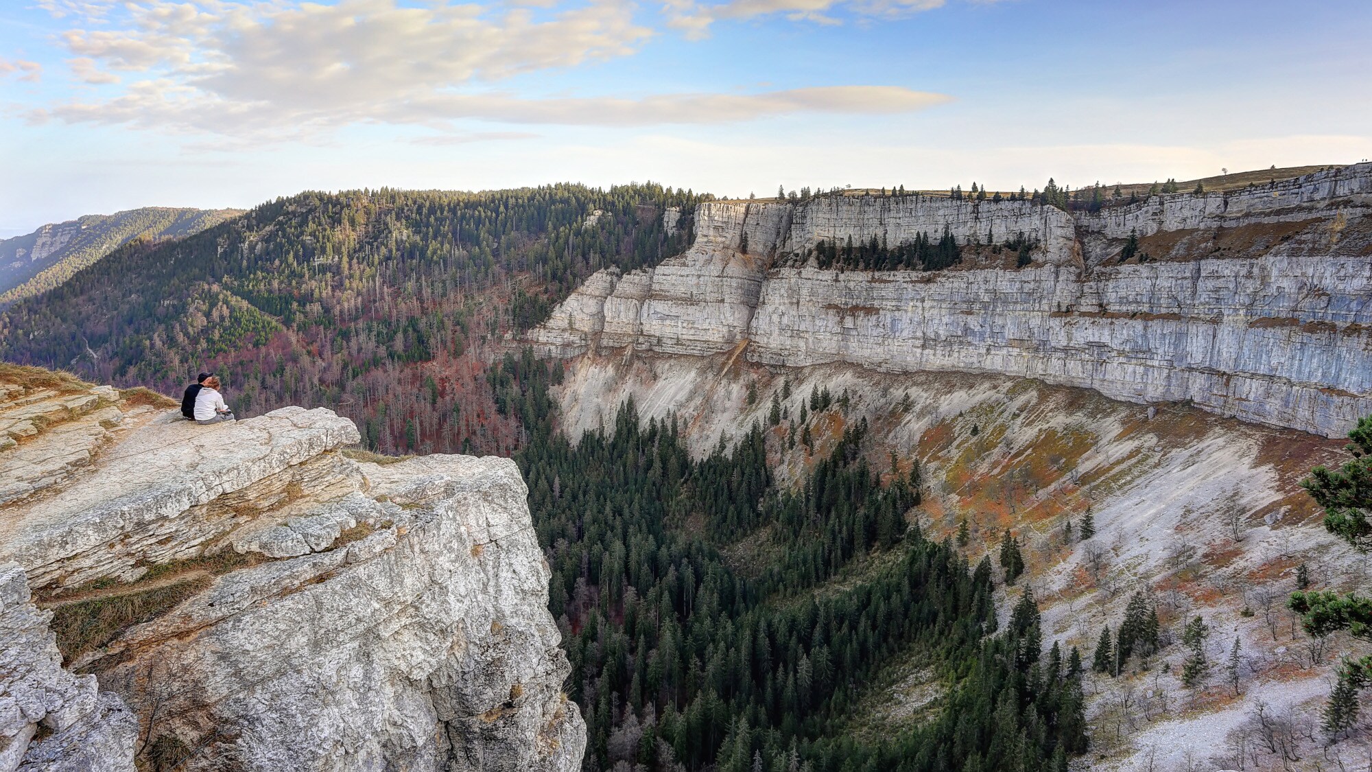 Zwei Personen sitzen auf einem Felsvorsprung vor einer gigantischen, senkrechten Felswand, umgeben von Waldlandschaft.