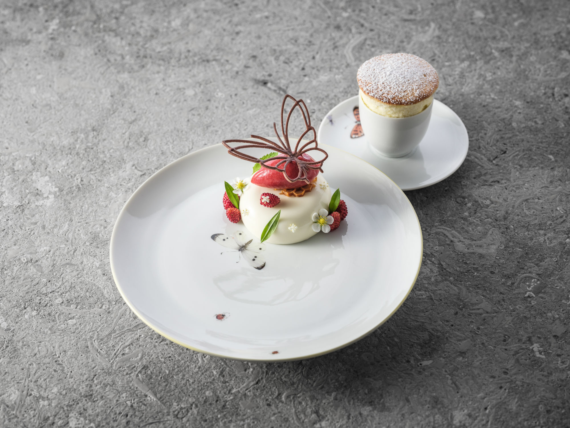 Stillleben zweier reduzierten, kunstvoll angerichteten Süßspeisen auf schlichten weißen Tellern auf anthrazitfarbenem Untergrund.