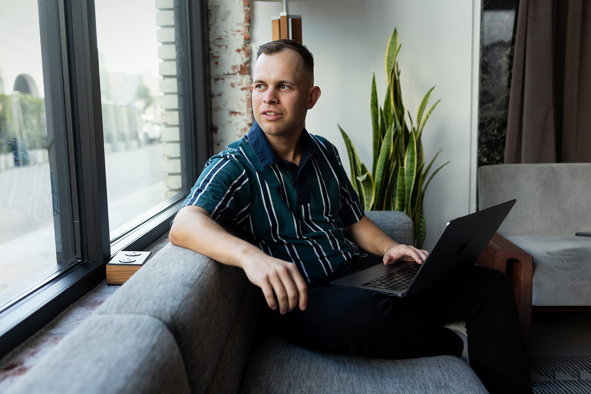 Business Stories: Von Business-Talenten für Business-Talente Benjamin Diedering sitzt mit Laptop auf dem Sofa und schaut aus dem Fenster.