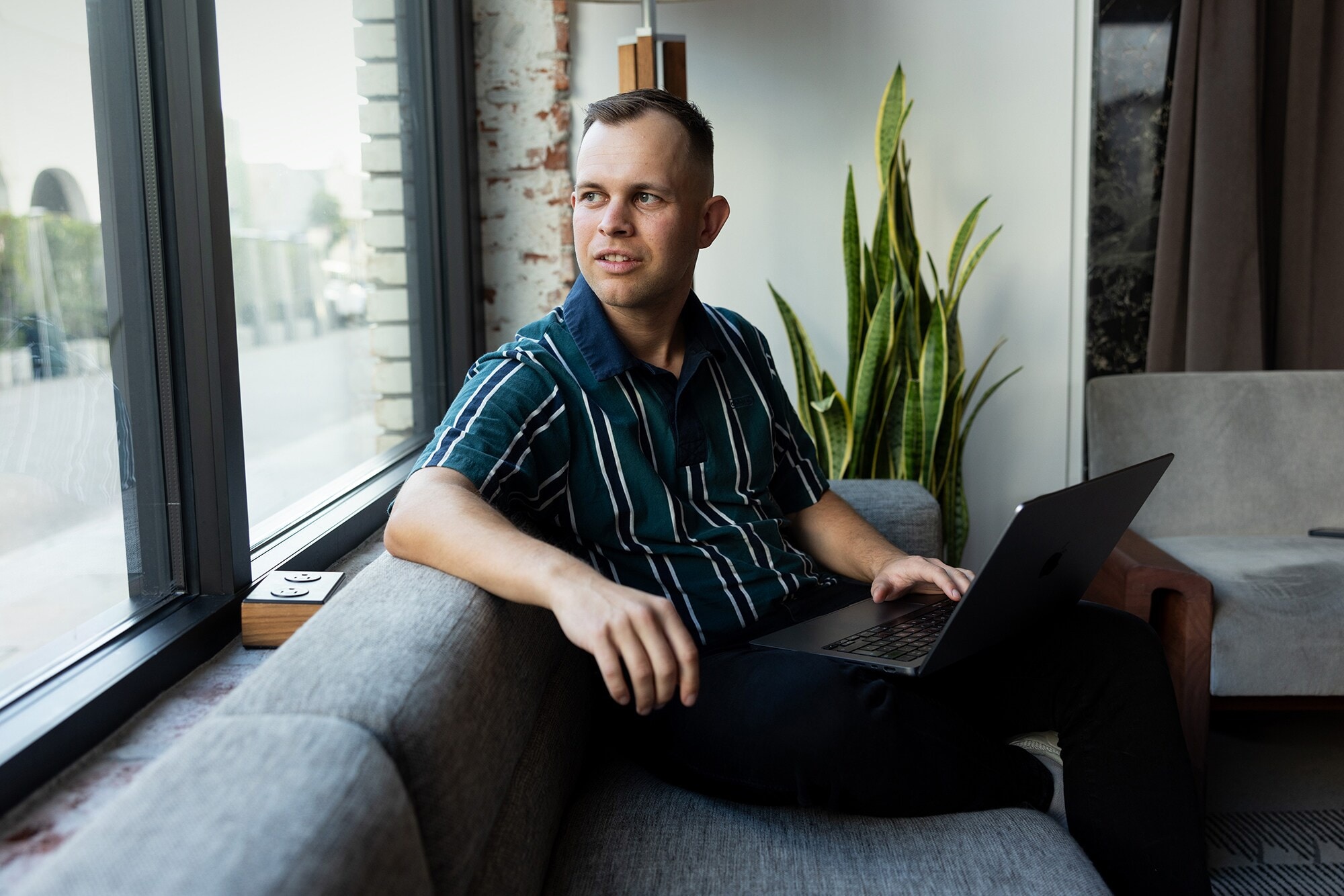 Business Stories: Von Business-Talenten für Business-Talente Benjamin Diedering sitzt mit Laptop auf dem Sofa und schaut aus dem Fenster.