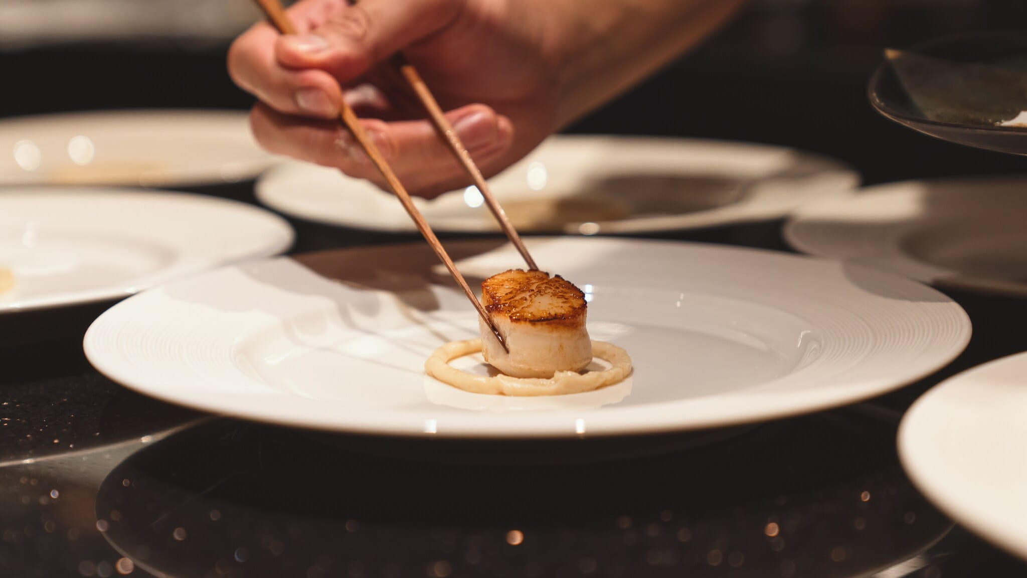Eine Person hält zwei Stäbchen an eine angebratene Jakobsmuschel auf einem weißen Teller. Eine Person hält zwei Stäbchen an eine angebratene Jakobsmuschel auf einem weißen Teller.