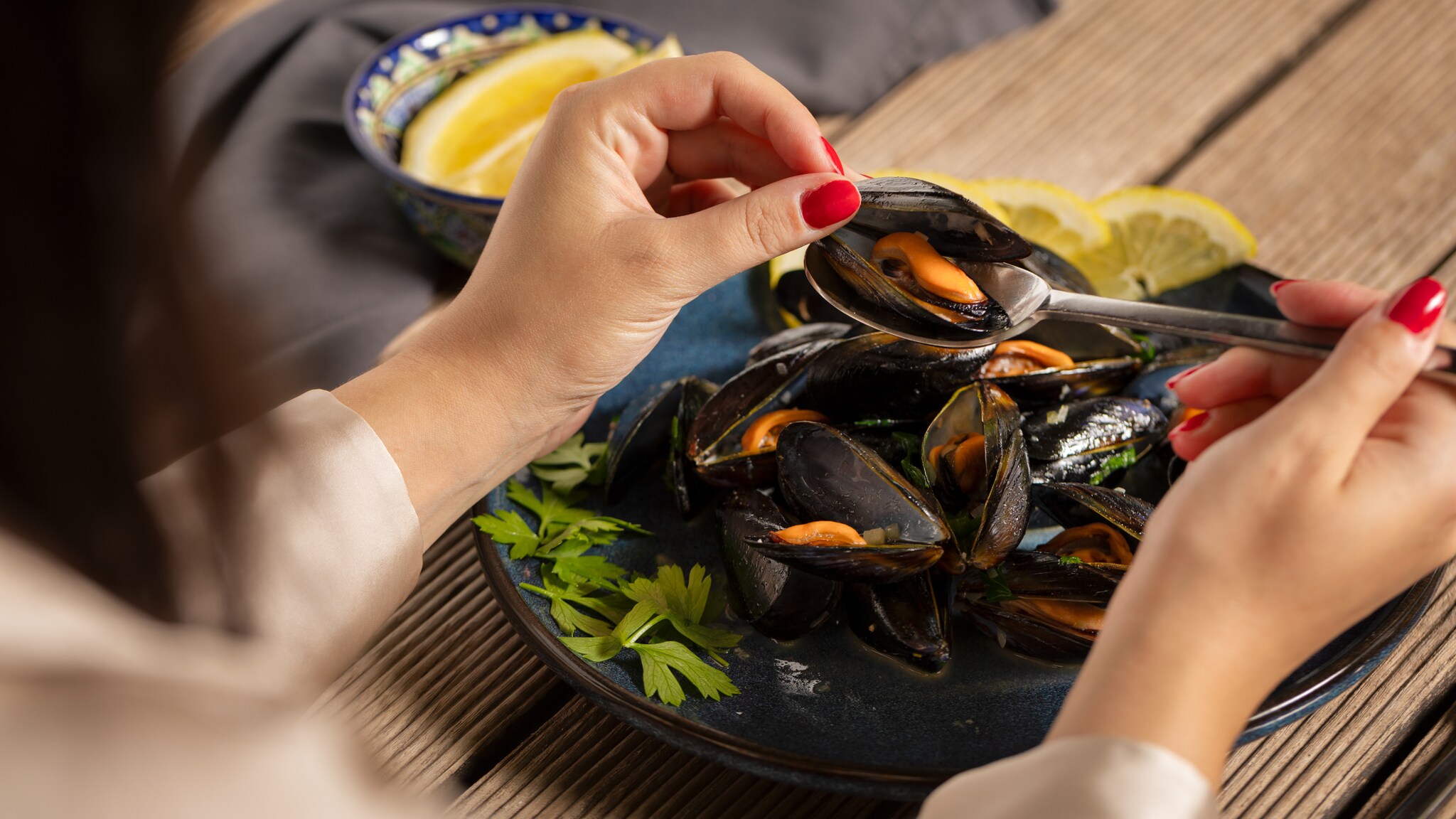 Eine Person vor einem Teller mit Muscheln hält mit einer Hand eine geöffnete Muschel und mit der anderen Hand einen Löffel, mit dem sie das Muschelinnere aus der Schale entfernt. Eine Person vor einem Teller mit Muscheln hält mit einer Hand eine geöffnete Muschel und mit der anderen Hand einen Löffel, mit dem sie das Muschelinnere aus der Schale entfernt.