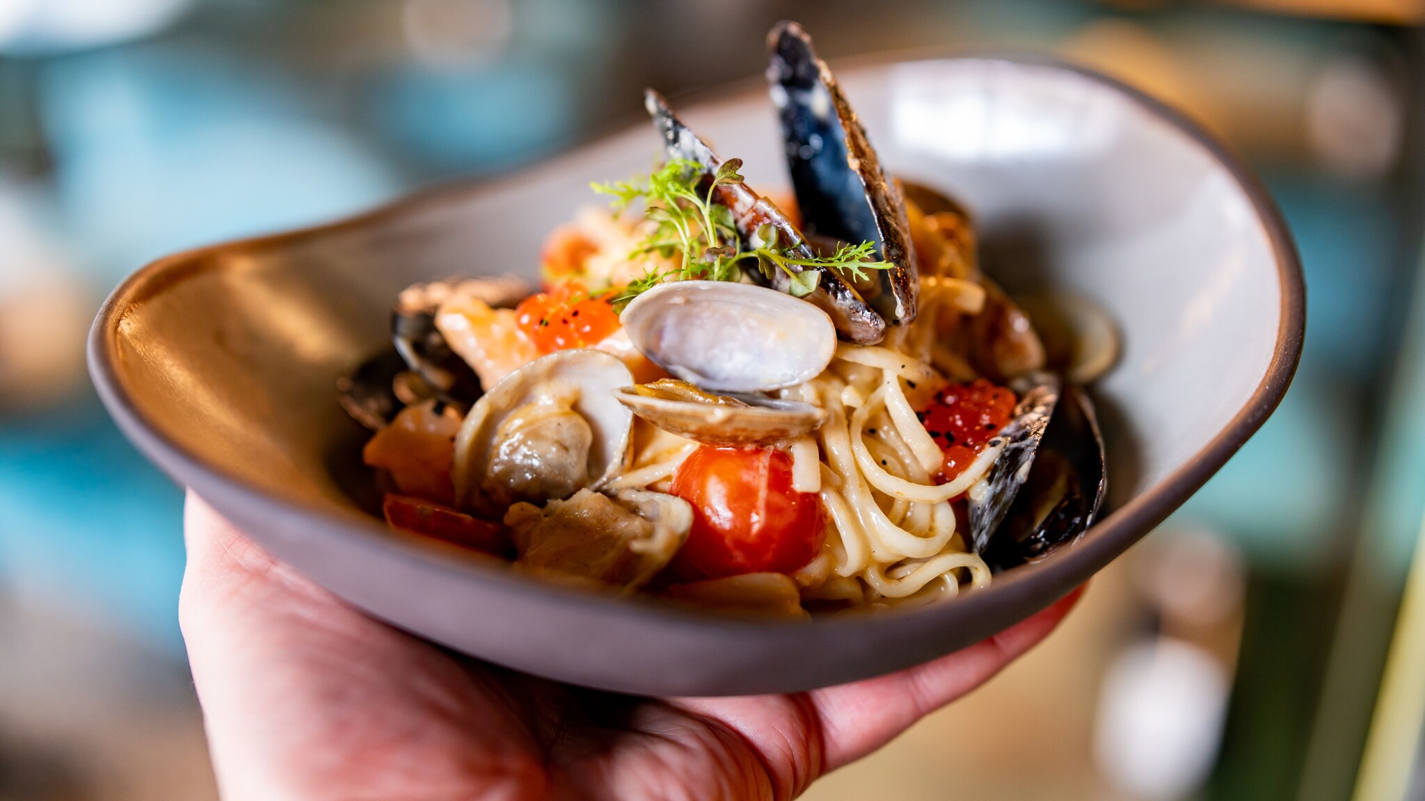 Eine Person hält eine Schale mit Pasta, die mit verschiedenen Muschelsorten und Tomaten angerichtet ist. Eine Person hält eine Schale mit Pasta, die mit verschiedenen Muschelsorten und Tomaten angerichtet ist.