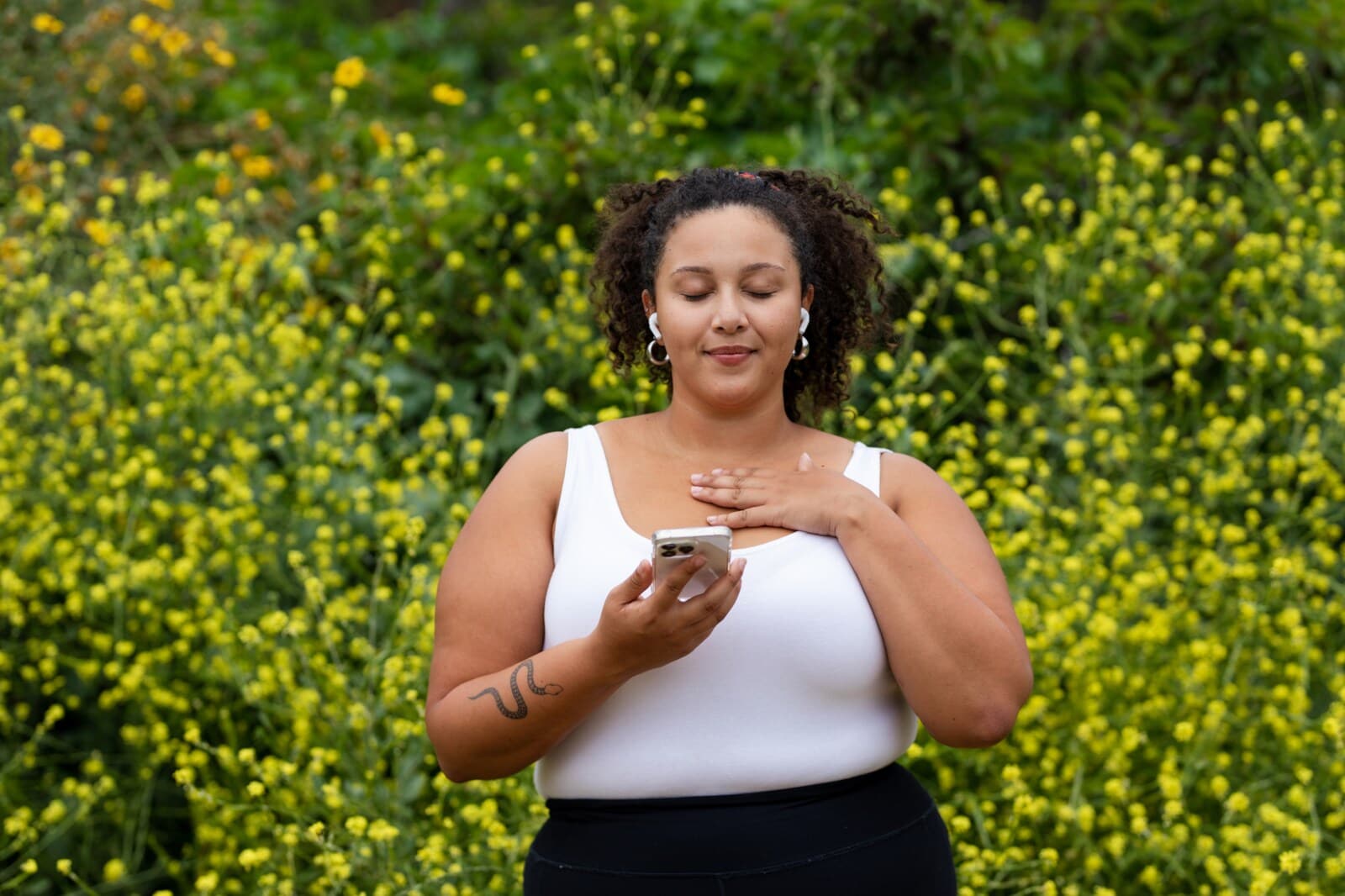 Eine Frau mit Smartphone in der Hand steht mit geschlossenen Augen in der Natur und meditiert.