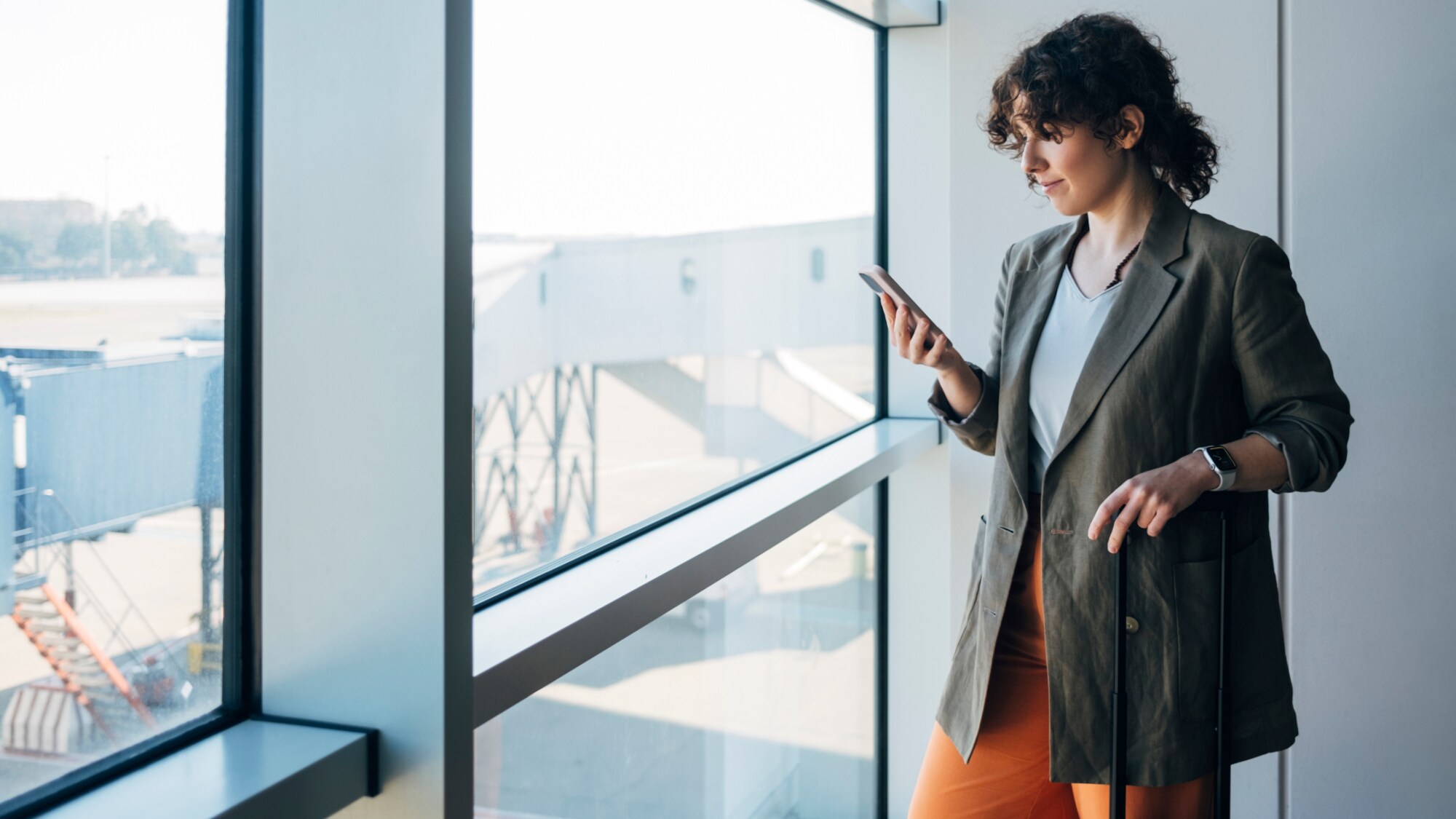 Eine Person, die am Fenster einer Flughafenhalle steht und auf ein Handy in ihrer Hand blickt – den anderen Arm hat sie auf einem Koffergriff abgelegt.