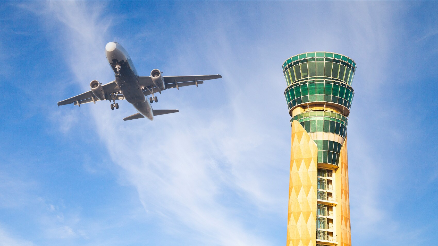 Untersicht eines Flugzeuges am blauen Himmel neben einem grün-gelben Tower.