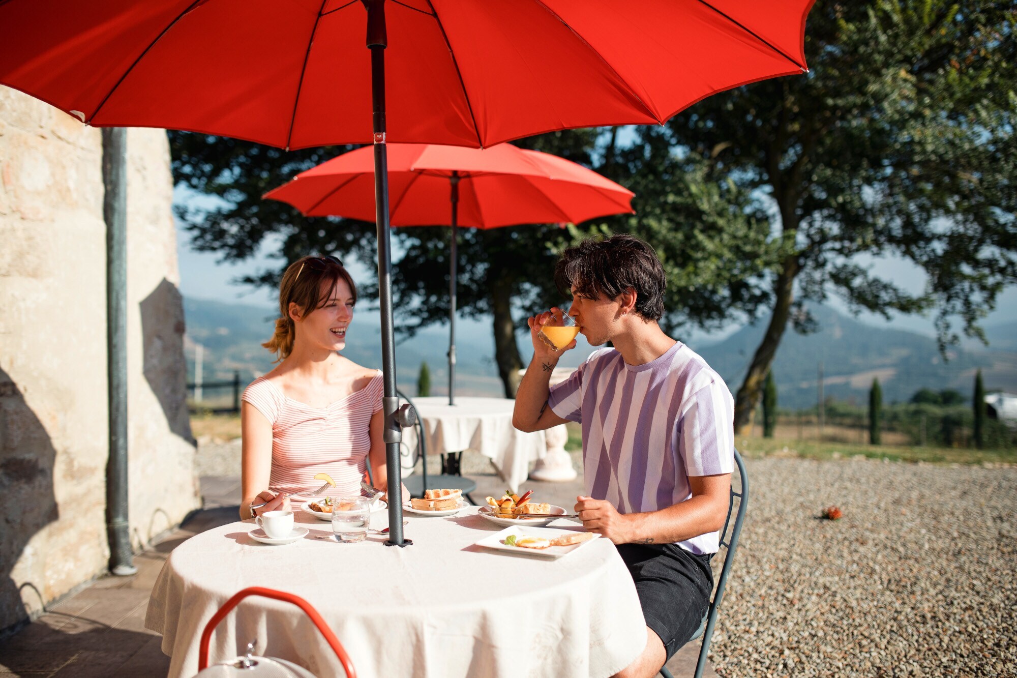 Ein Paar frühstückt auf einer Terrasse an einem Tisch bei Sonnenschein, im Hintergrund eine Hügellandschaft.